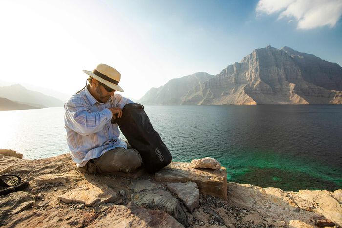 a man sitting on a rocky outcropping overlooking some water and reaching into his seal line dry bag