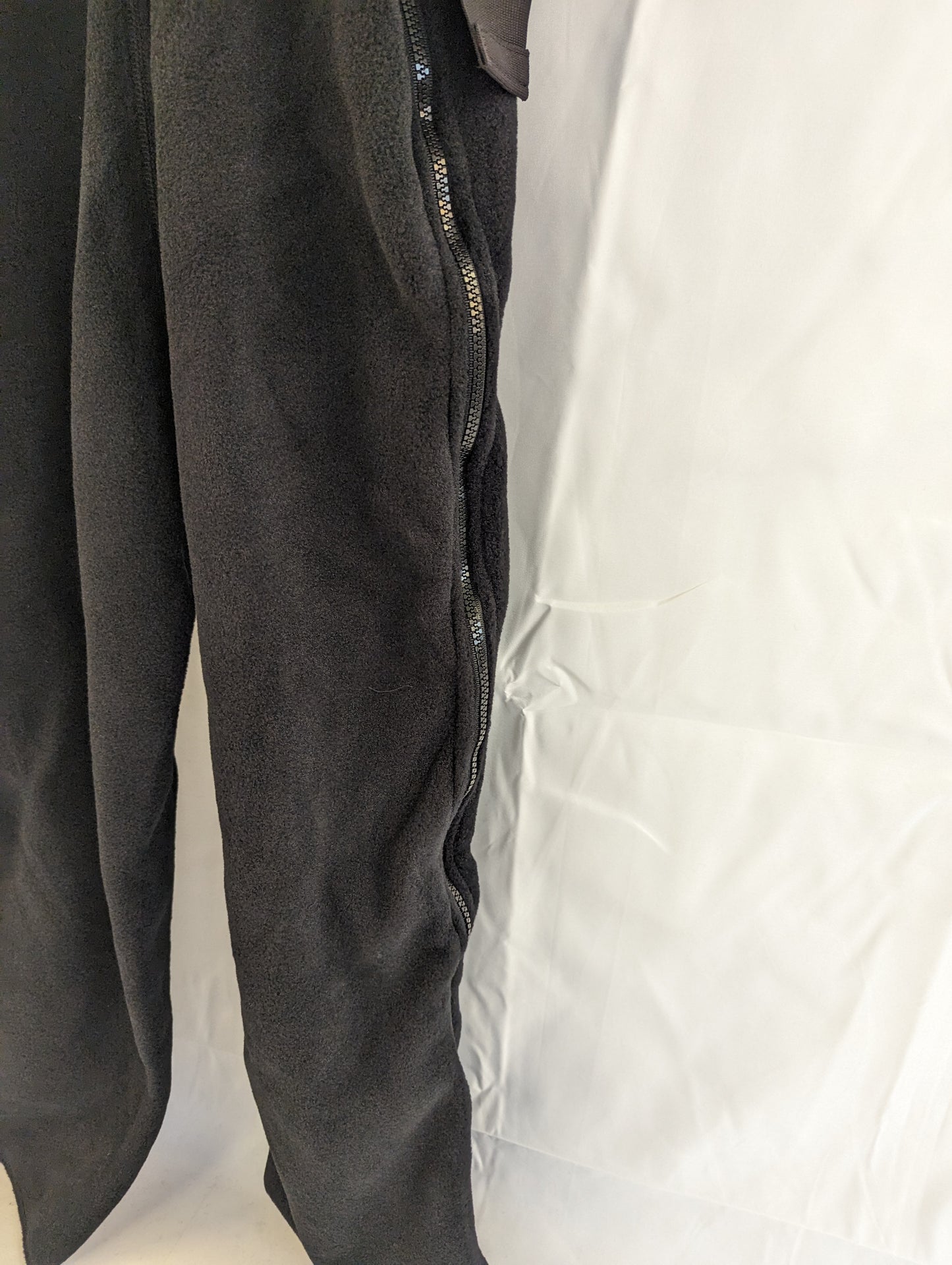 A close-up detail shot of the side zipper on the leg of the black Polartec fleece cold weather overalls, showing the zipper track and the texture of the fleece fabric.
