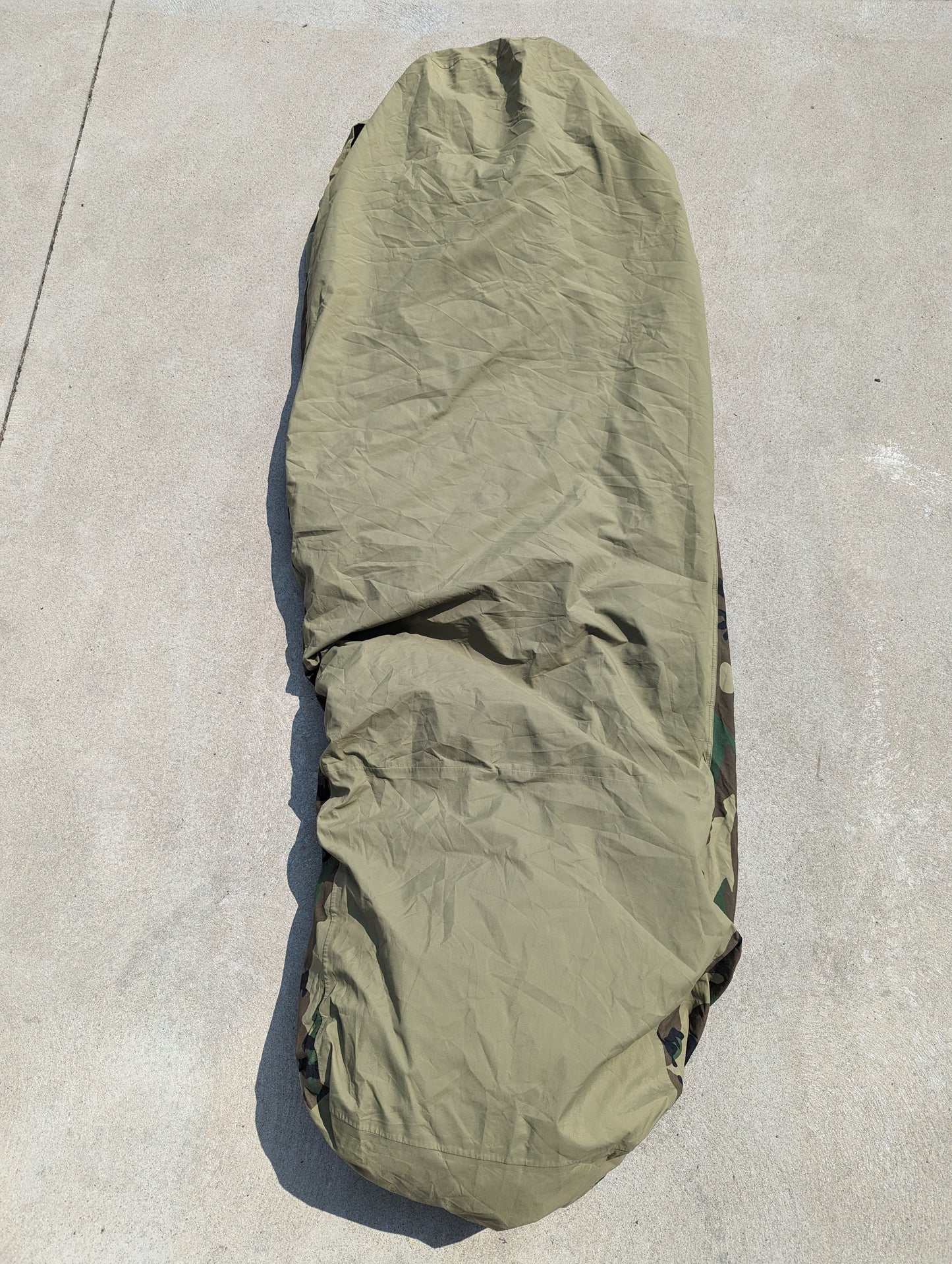 A full-length U.S. Military Surplus Modular Sleep System Bivy Cover in solid olive green, zipped closed and laid flat on a concrete surface, displaying its wrinkled, waterproof fabric.