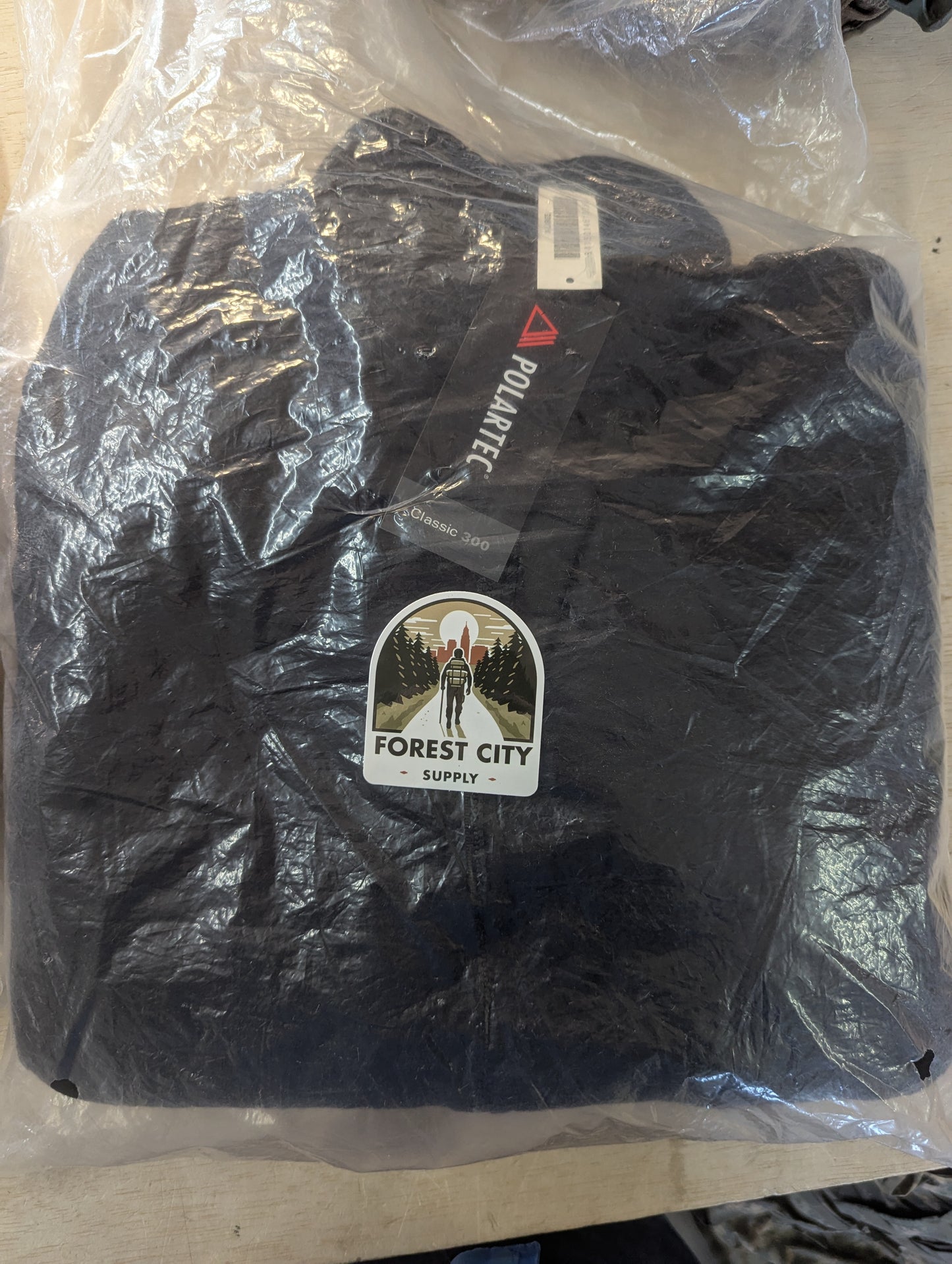 A brand new, packaged black Polartec fleece jacket sealed in clear plastic, featuring a 'FOREST CITY SUPPLY' logo sticker and visible manufacturer hang tags.