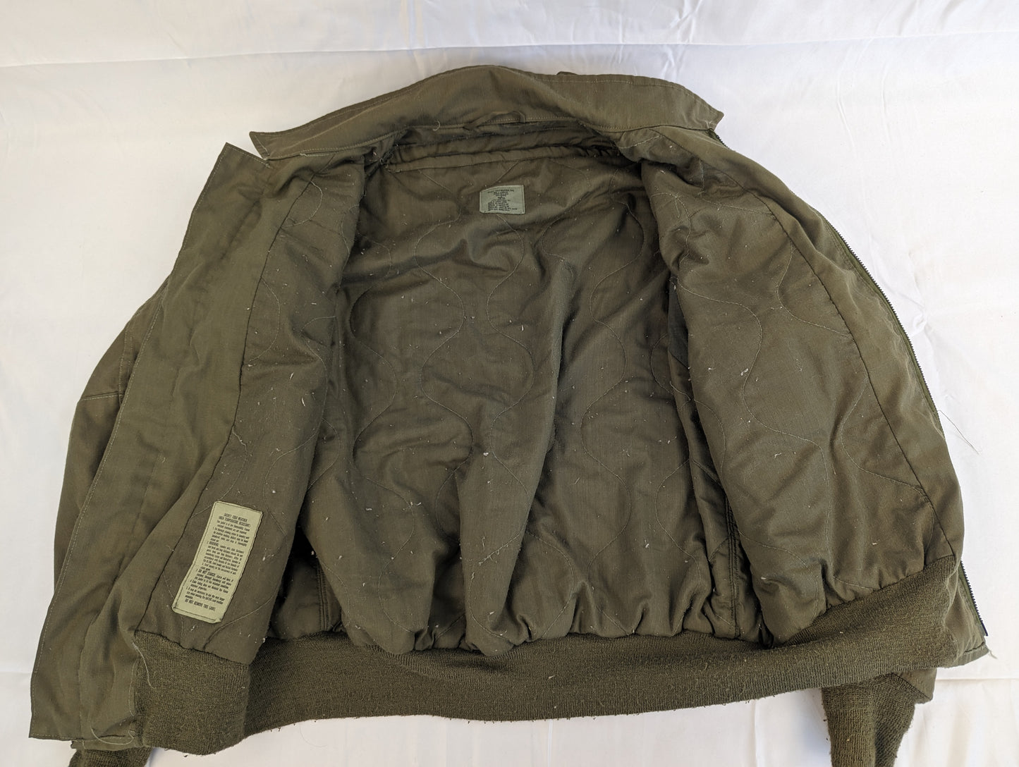 A back view of the U.S. Military CVC jacket turned inside-out, displaying the quilted olive green liner, internal tags, and the ribbed collar and waistband.