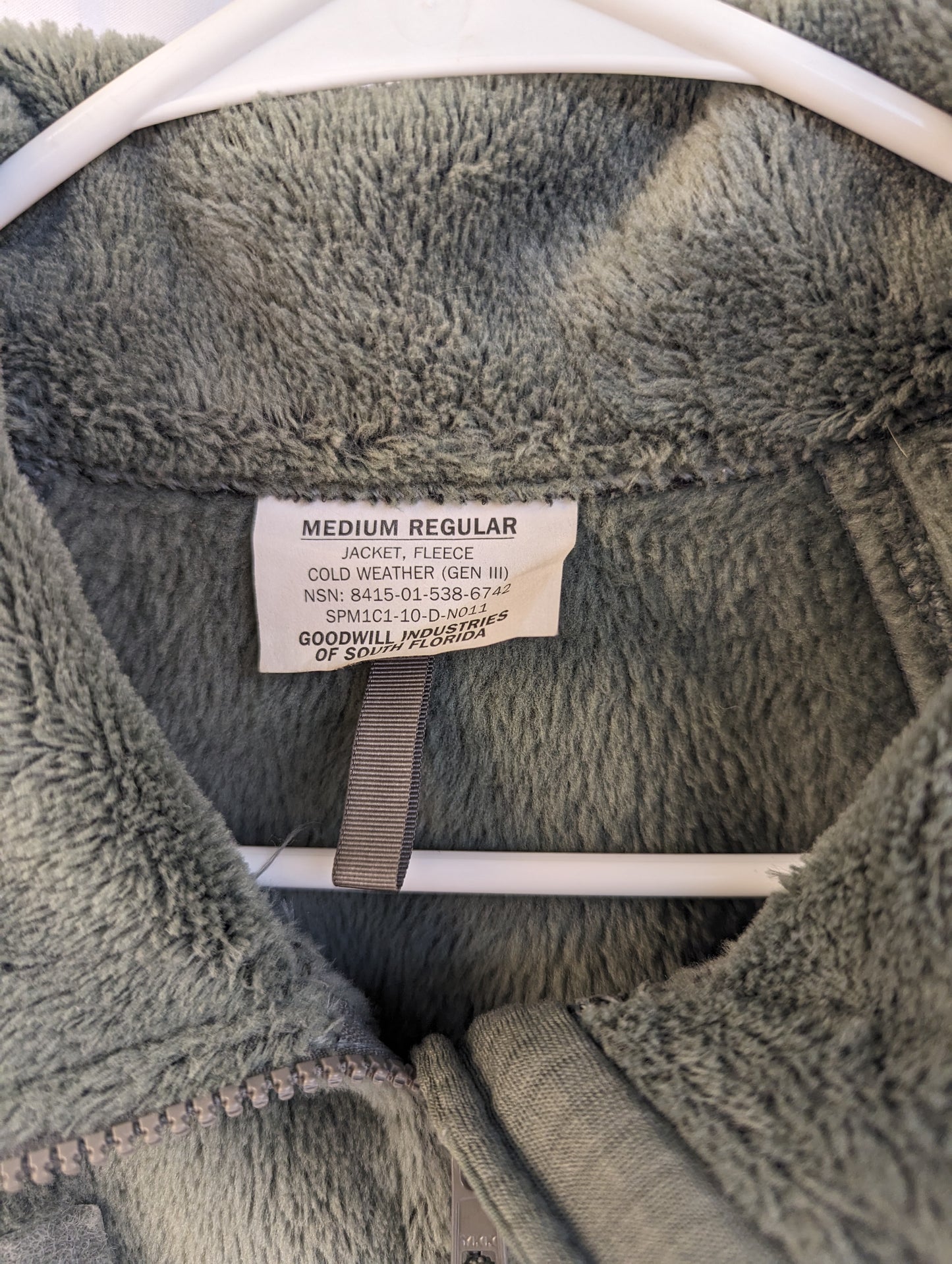 A close-up photograph of the official U.S. Military issue tag sewn into the green fleece jacket, clearly displaying the size 'MEDIUM REGULAR', the National Stock Number 8415-01-538-6794, and manufacturer 'Goodwill Industries'.