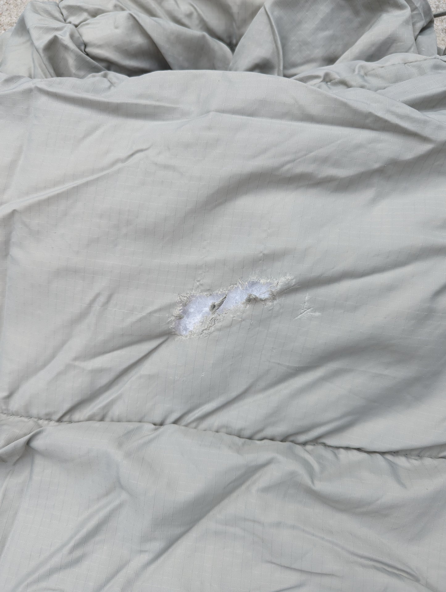 A close-up photograph of the quilted foliage green fabric of a military sleeping bag, highlighting a tear with white insulation visible, documenting damage.