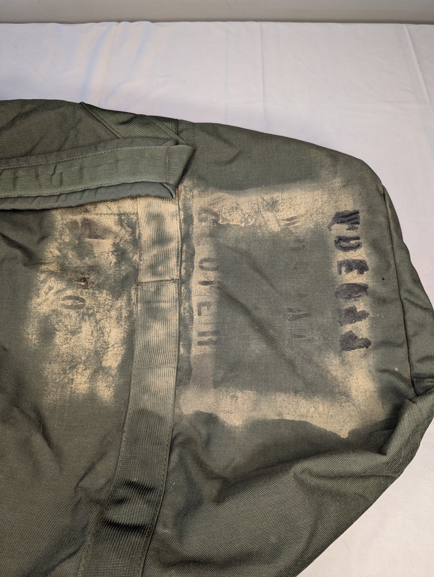 A close-up detail shot of the olive green U.S. Military Improved Duffel Bag fabric, the heavy wear, and a large, faded stenciled government marking on the side.