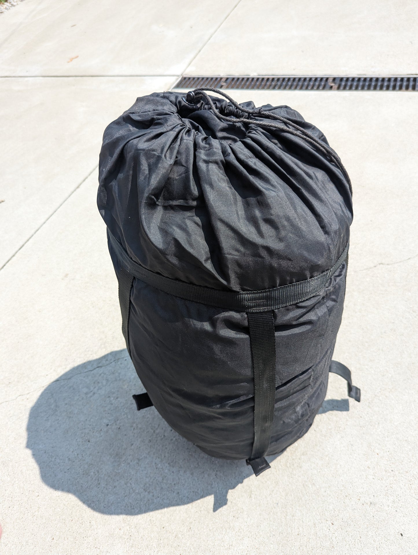 A large, tightly compressed U.S. Military Surplus Compression Stuff Sack in black, standing upright on a concrete surface, featuring a drawstring closure and heavy webbing compression straps.