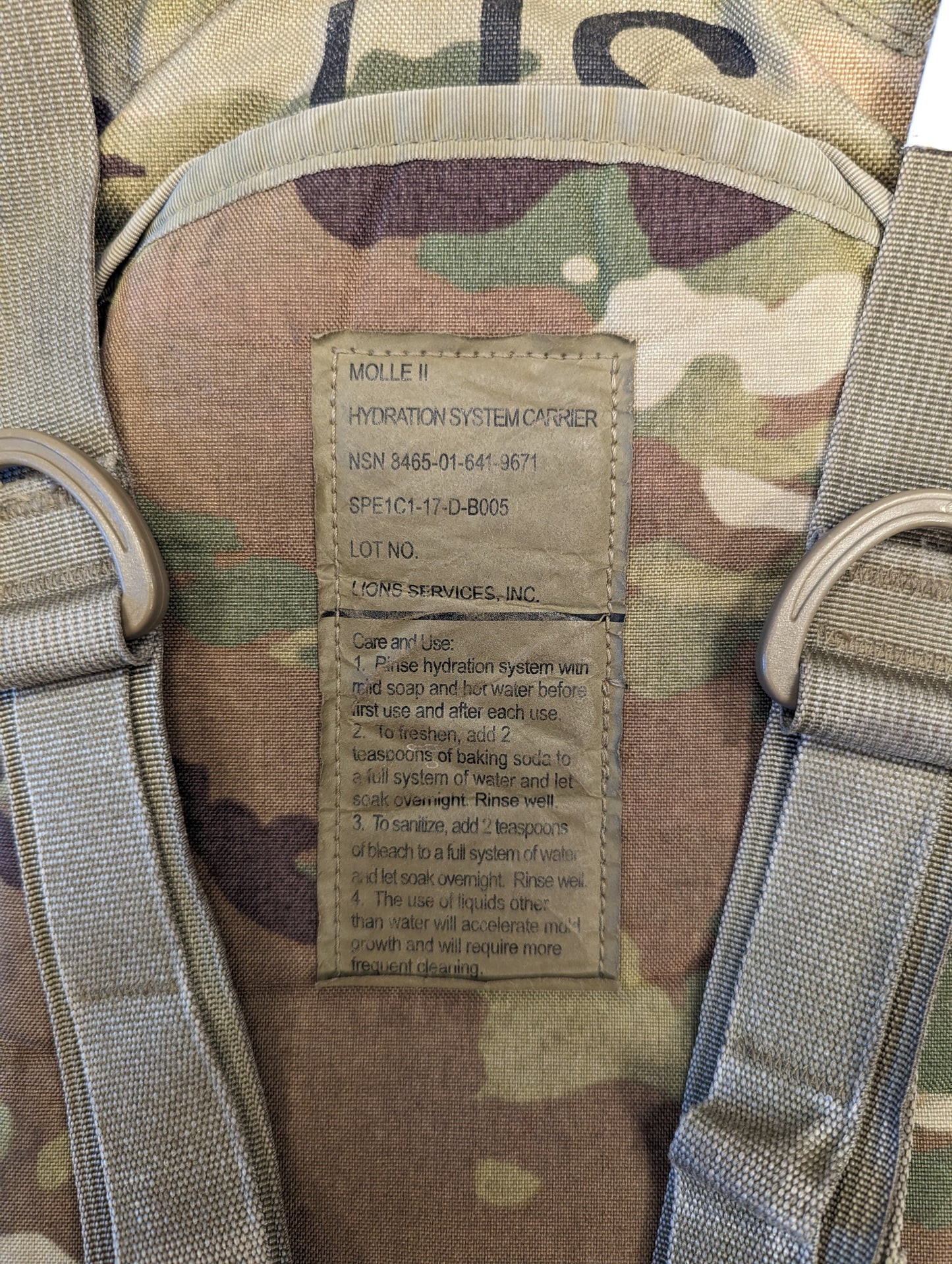 A close-up photograph of the official U.S. Military issue tag sewn into the OCP/Multicam Hydration Carrier, detailing the product name, NSN, Camelbak manufacturer, and cleaning instructions.