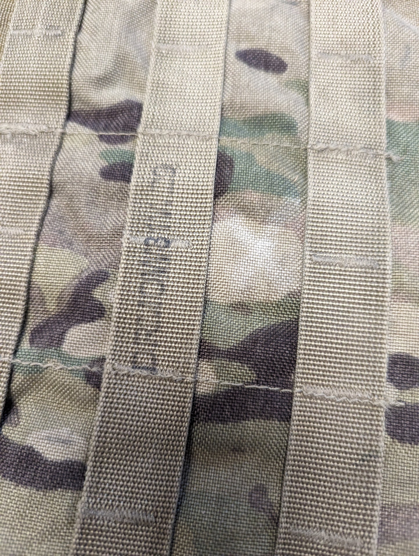 A close-up detail shot of OCP/Multicam camouflage PALS/MOLLE webbing, showing the horizontal and vertical strap construction, with the letters 'US' faintly marked on one of the straps.