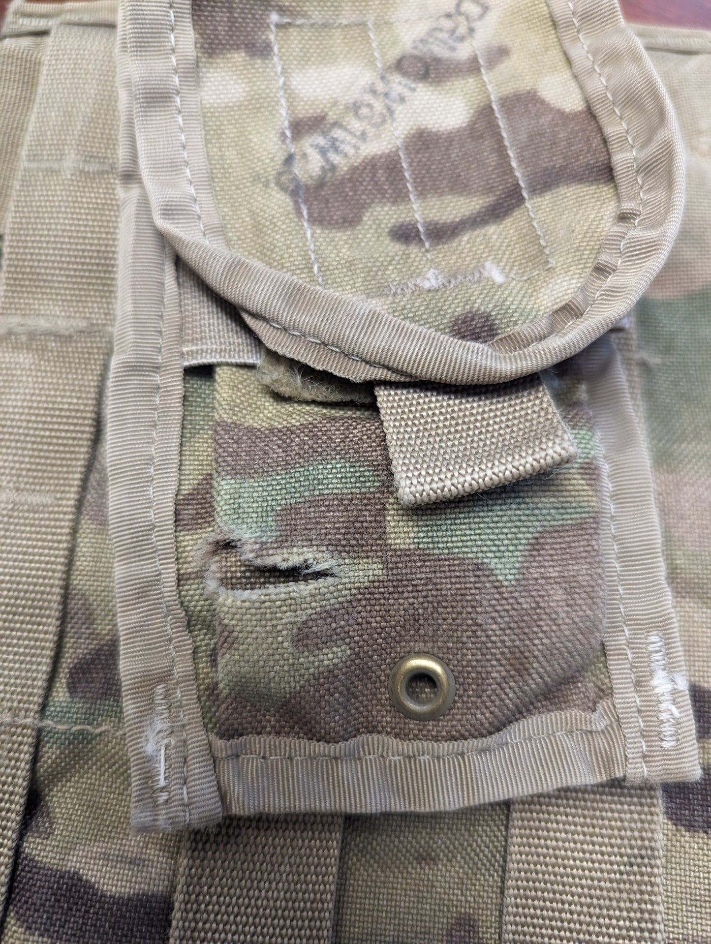 An extreme close-up detail shot of an OCP/Multicam tactical pouch flap, highlighting a tear or hole in the fabric just above a brass drainage grommet, documenting a specific surplus defect.