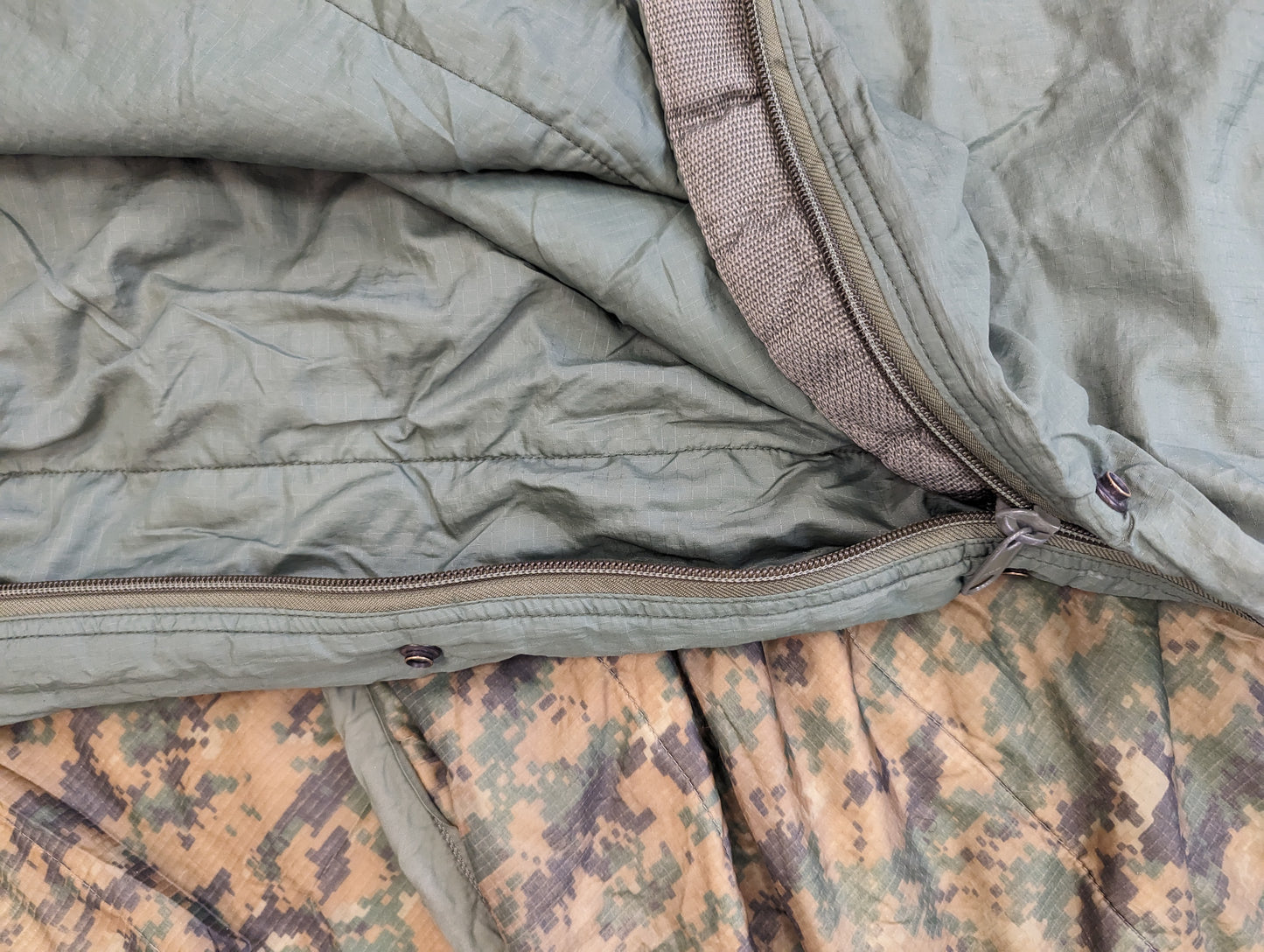 A close-up view of the light olive green U.S. Military Surplus Patrol Sleeping Bag, highlighting the zipper closure and quilted material, resting on an OCP camouflage background.
