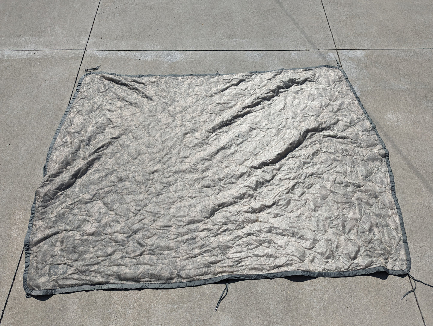 A full-length U.S. Military Surplus Poncho Liner (Woobie) in a muted gray/ACU camouflage, laid flat on a concrete surface, displaying its wrinkled texture, edge binding, and attachment tie cords.