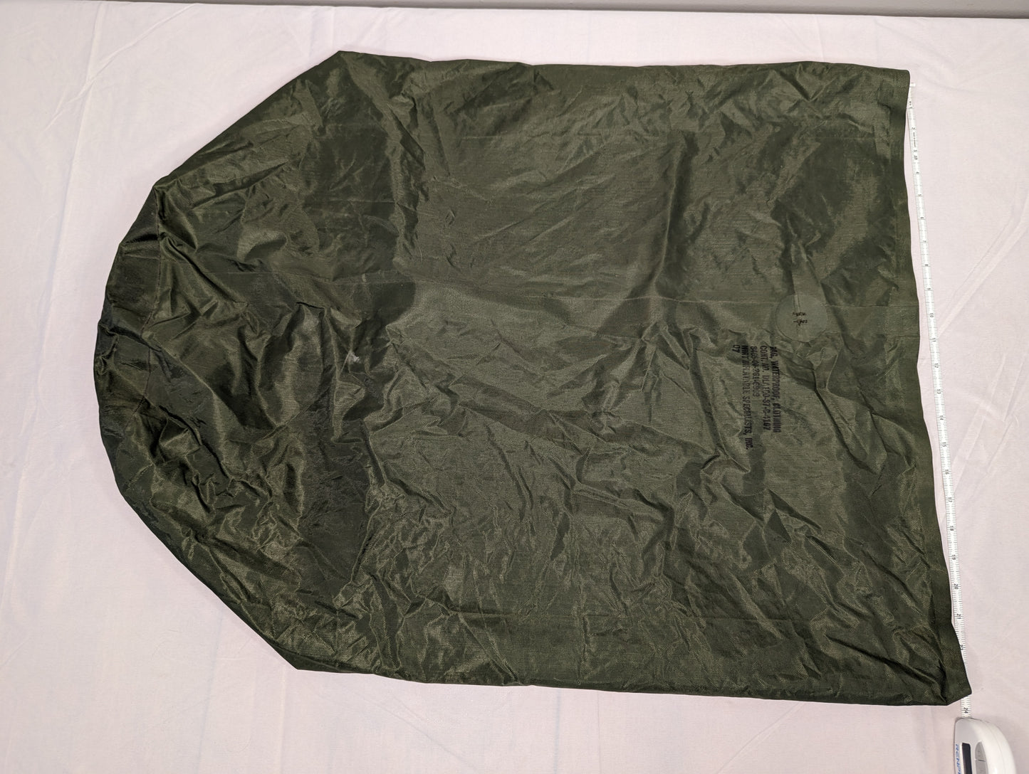 A large, fully unfolded U.S. Military Waterproof Clothing Bag in dark olive green, laid flat on a white surface, displaying its ripstop texture and large dimensions with a measuring tape along the edge.