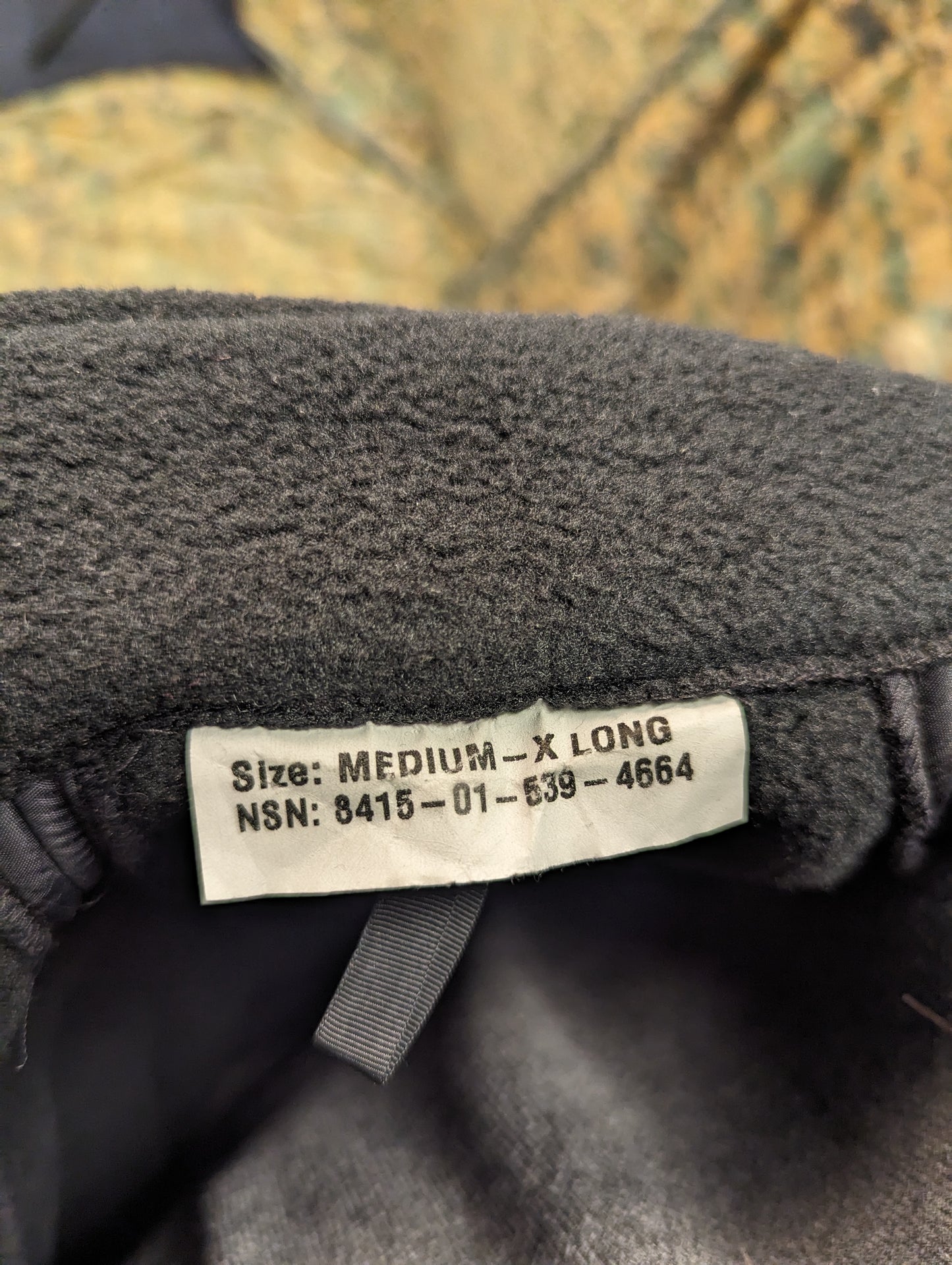 A close-up photograph of the U.S. Navy Parka Liner sizing tag, clearly displaying the size 'MEDIUM - X LONG' and the National Stock Number (NSN).