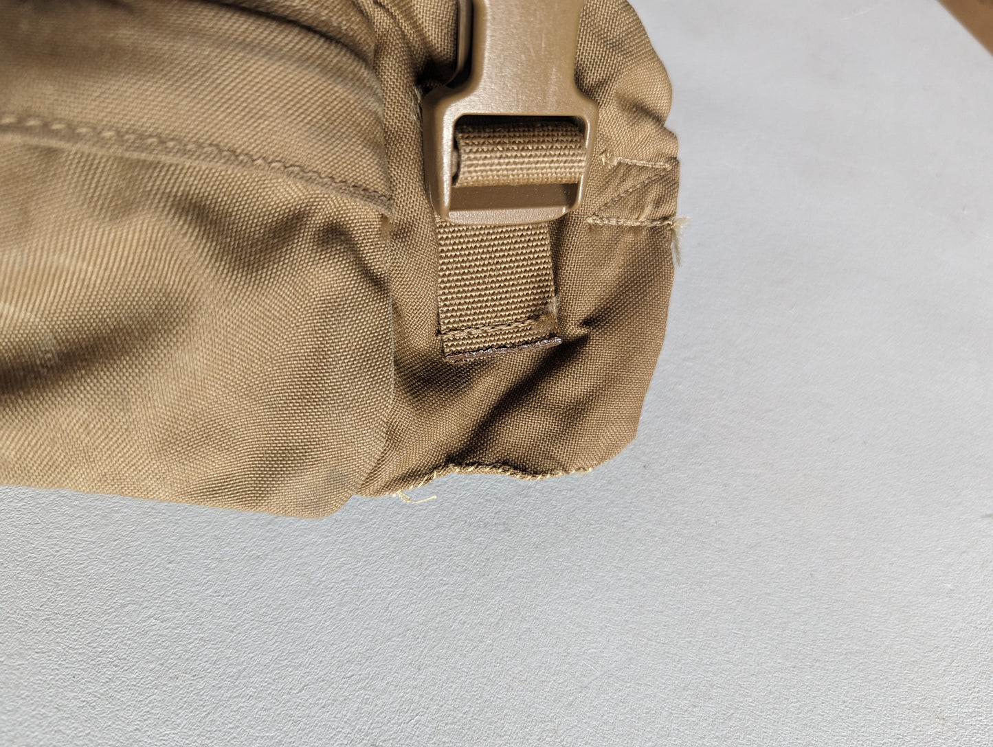 An extreme close-up detail shot of a Coyote Brown tactical pack, showing a tan plastic quick-release buckle attached to webbing, with the fabric heavily frayed and damaged at the bottom edge.