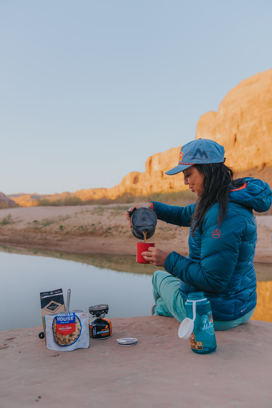 Person sitting by a lake with camping gear, wearing a blue jacket and cap.