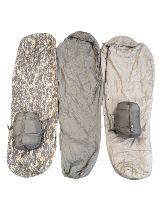 A collection of camouflage-patterned modular sleep system components, including a sleeping bag and a bivy cover, laid out on a black background.