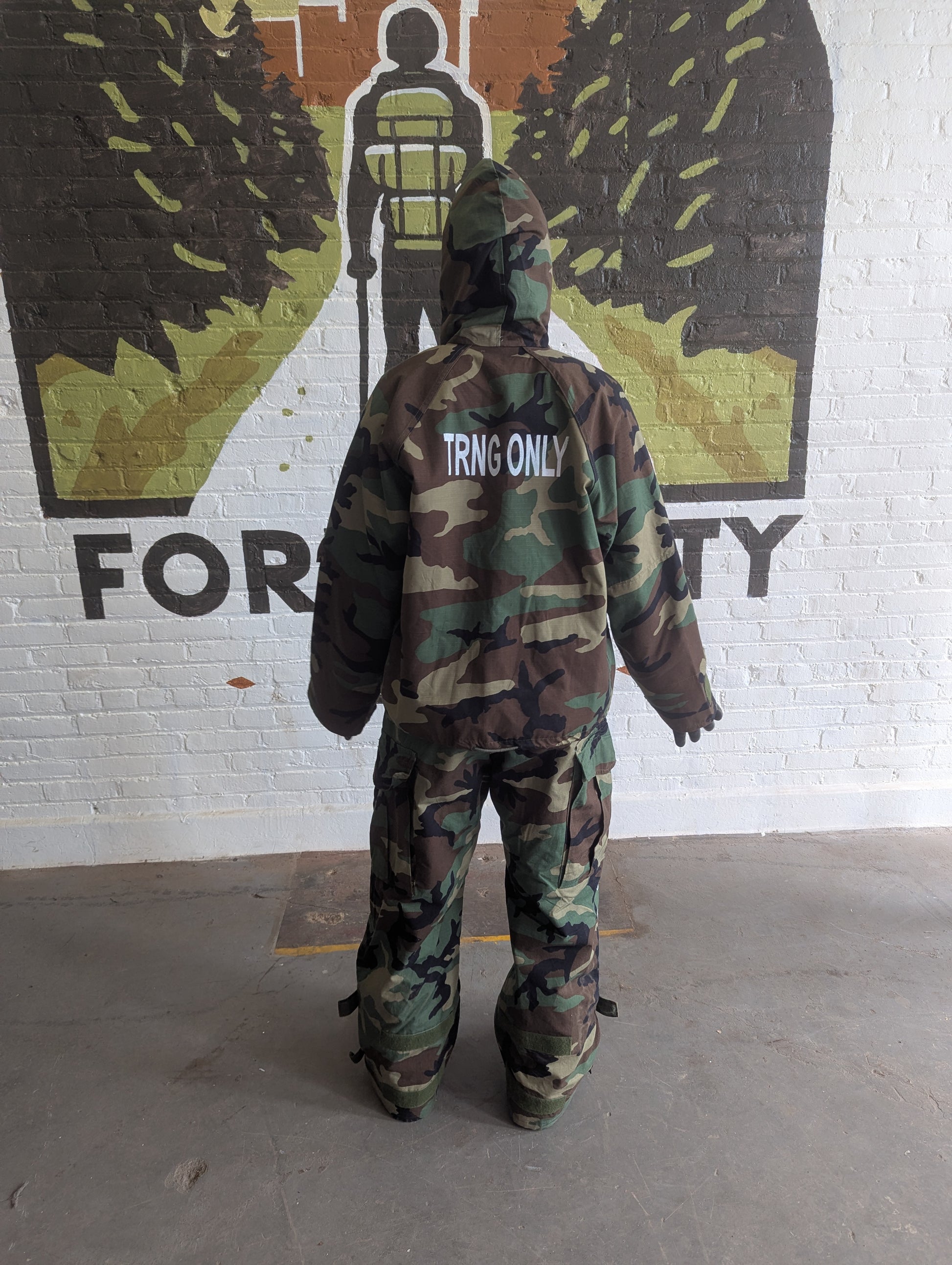 Person wearing a camouflage snowsuit with 'TRNG ONLY' text, standing in front of a mural on a white brick wall.