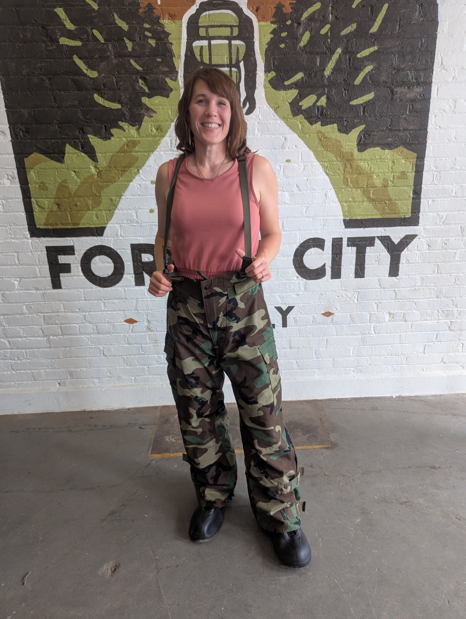 Person wearing a pink tank top and camouflage pants standing in front of a mural with 'For City' text.