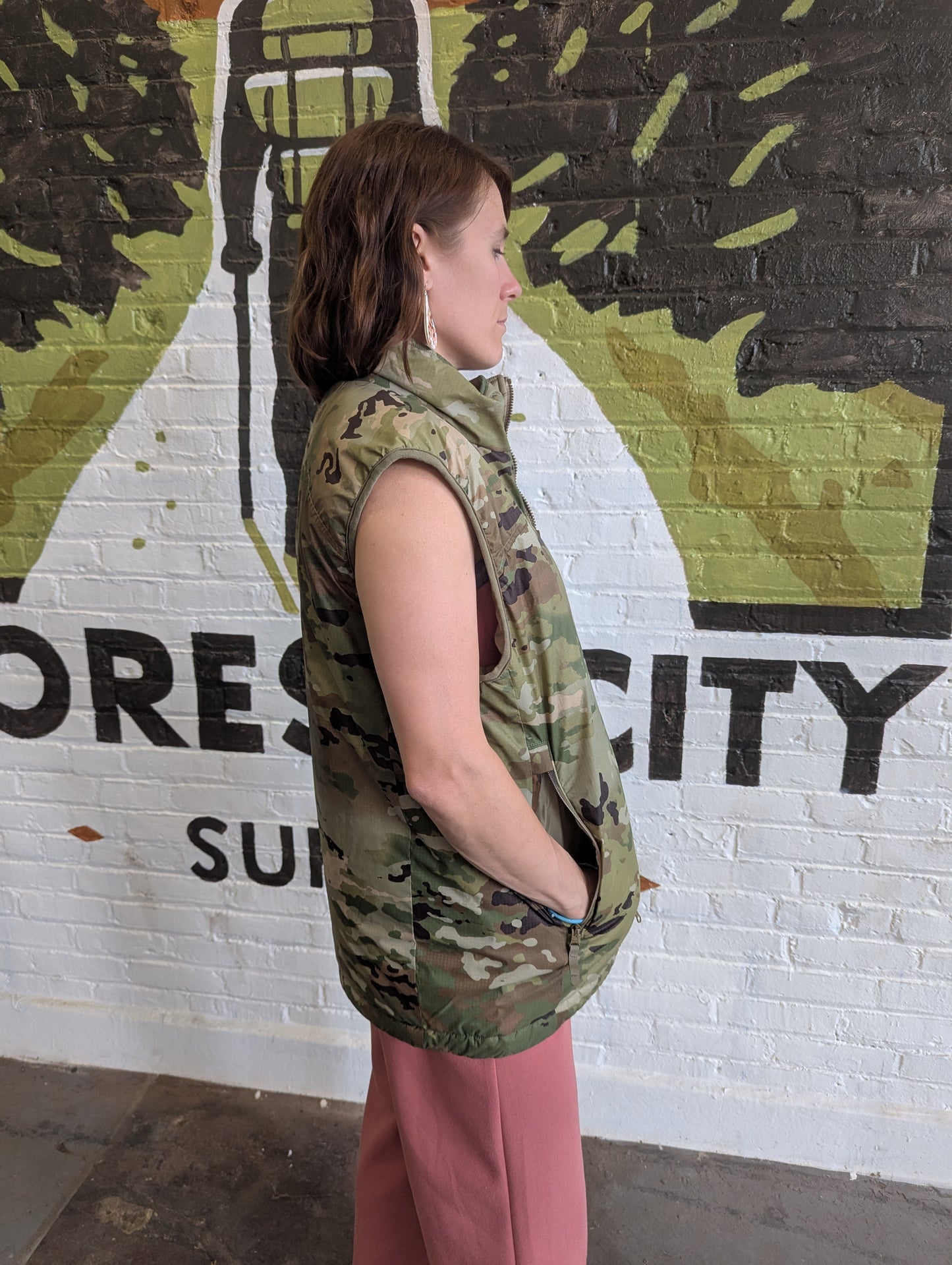 Woman wearing a camouflage vest standing in front of a mural with text on a wall.