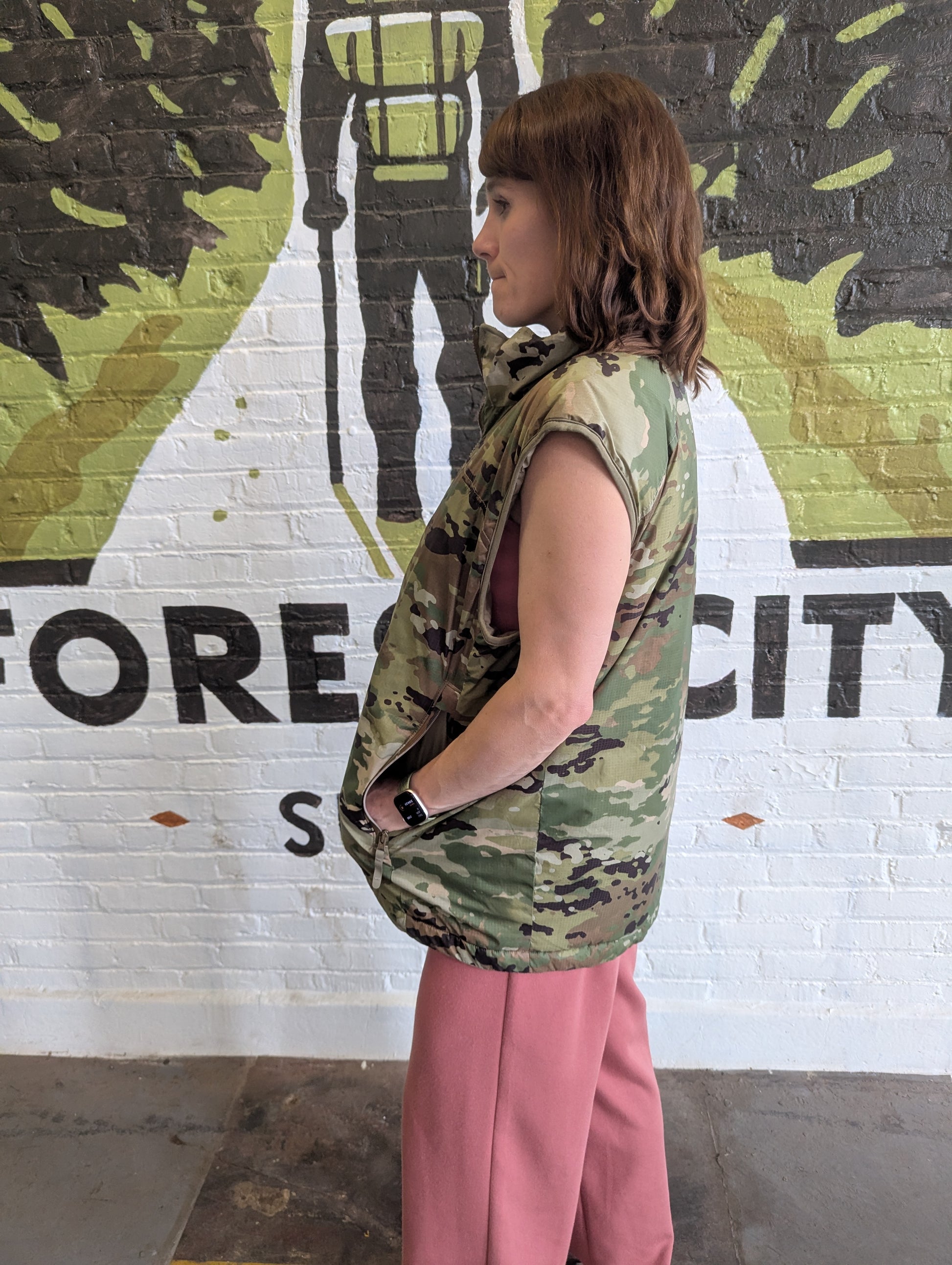Person wearing a camouflage vest and pink pants standing in front of a mural with text.