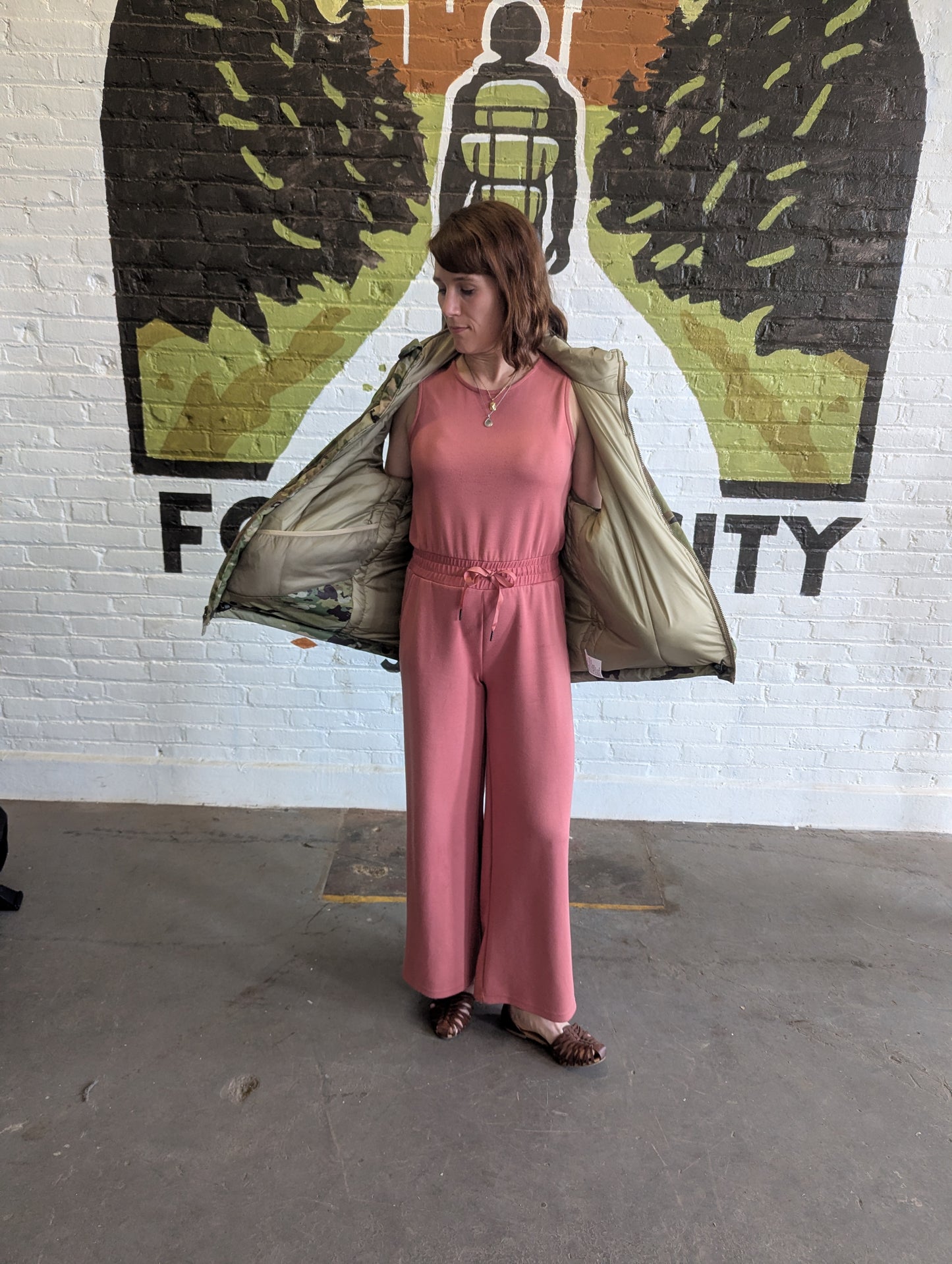 Person wearing a pink outfit with a beige jacket in front of a mural on a white brick wall.
