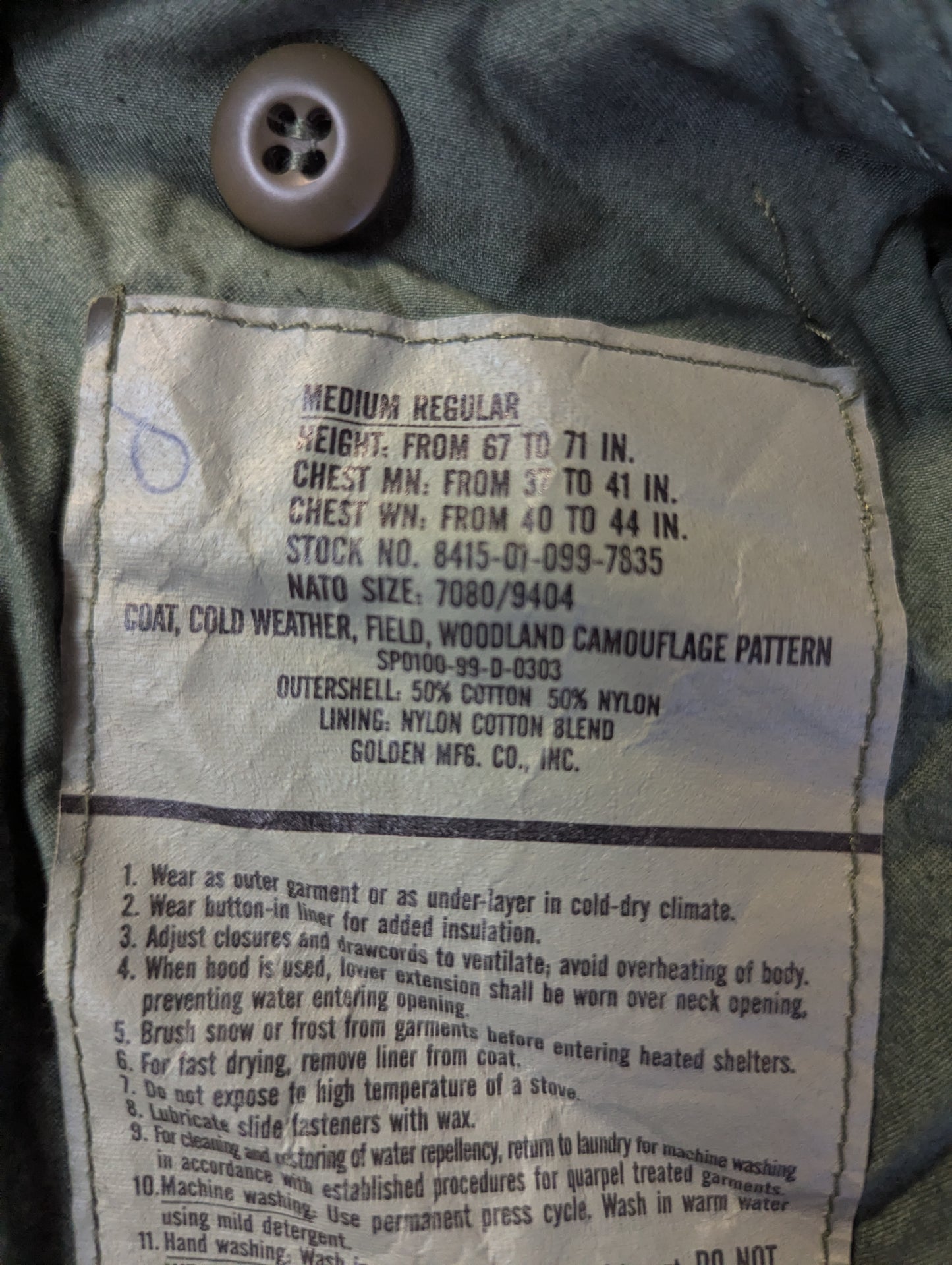Close-up of a faded beige cotton label sewn onto dark gray or black denim fabric, showing printed text and a buttonhole hole.