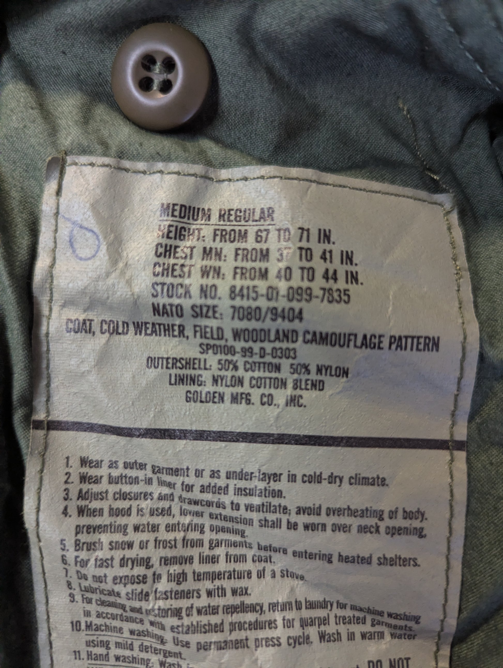 Close-up of a faded beige cotton label sewn onto dark gray or black denim fabric, showing printed text and a buttonhole hole.