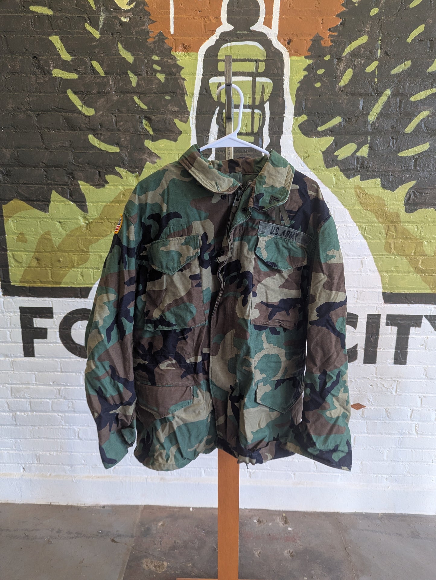A vintage woodland camouflage utility jacket hanging on a white hanger from a wooden stand, set against a white brick wall with a colorful graffiti mural.