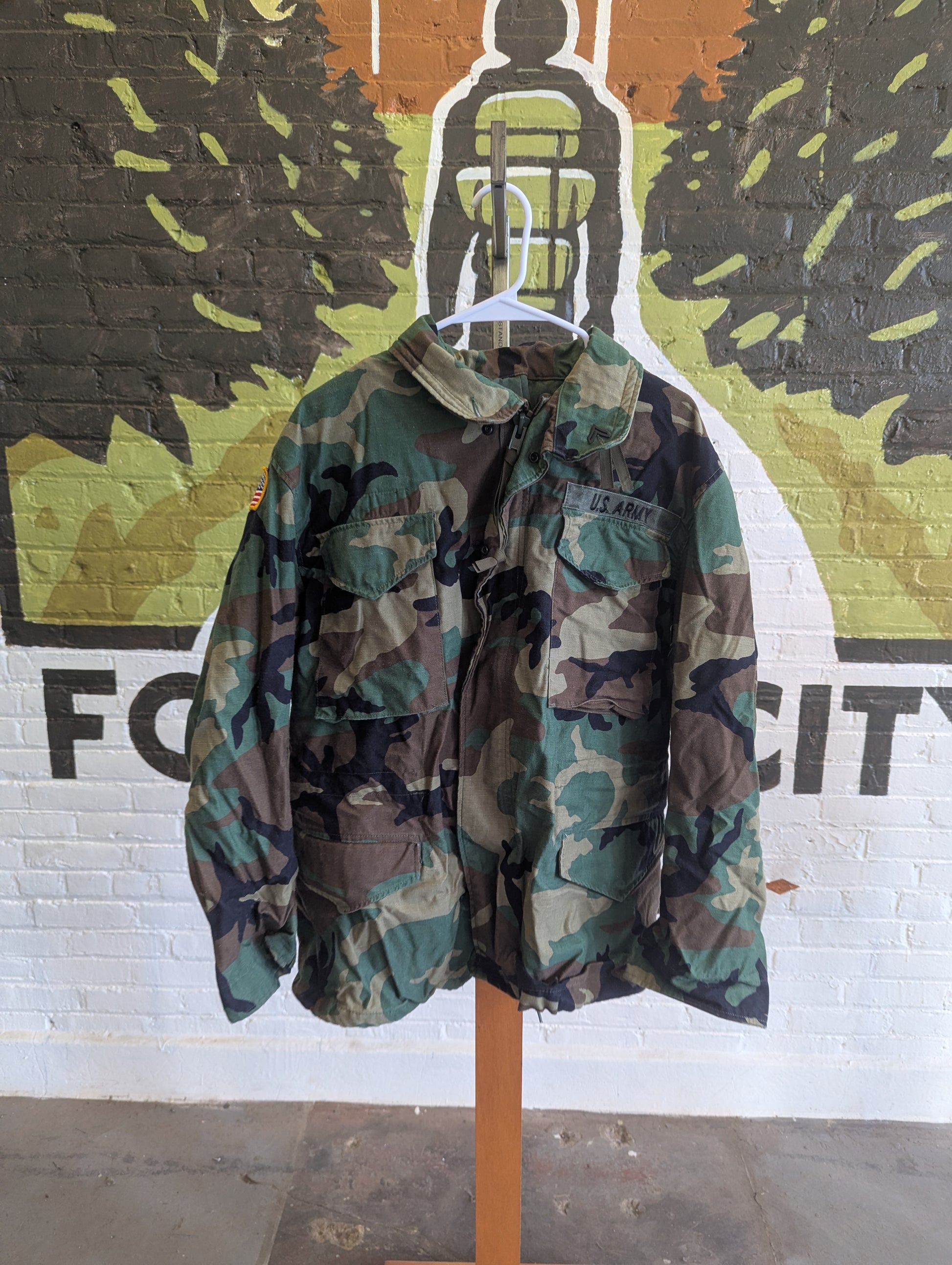 A vintage woodland camouflage utility jacket hanging on a white hanger from a wooden stand, set against a white brick wall with a colorful graffiti mural.