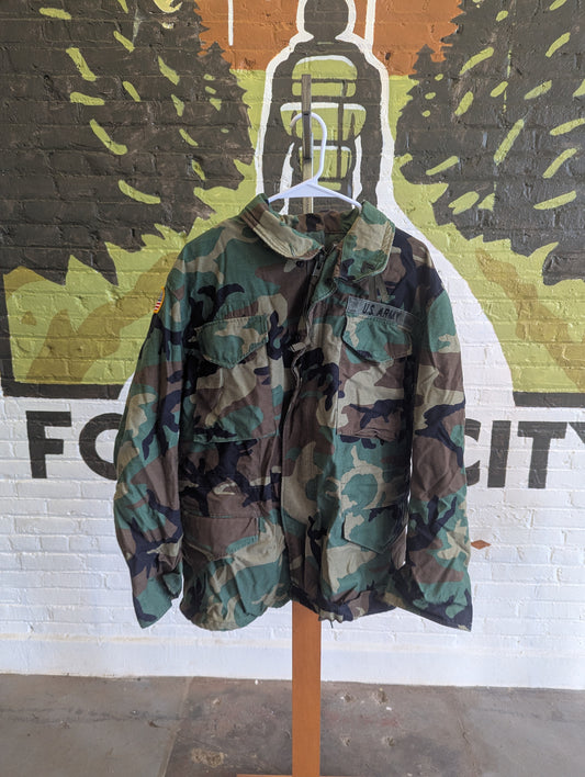 A vintage woodland camouflage utility jacket hanging on a white hanger from a wooden stand, set against a white brick wall with a colorful graffiti mural.