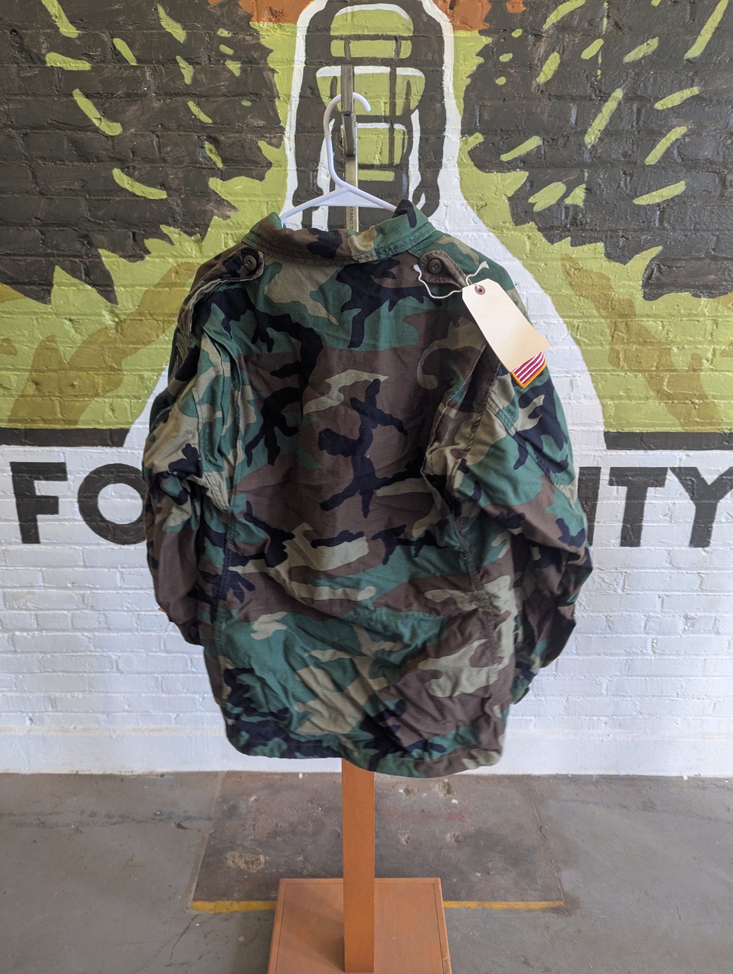 A camouflage hooded jacket displayed on a wooden hanger stand against a painted brick wall with a large mural.