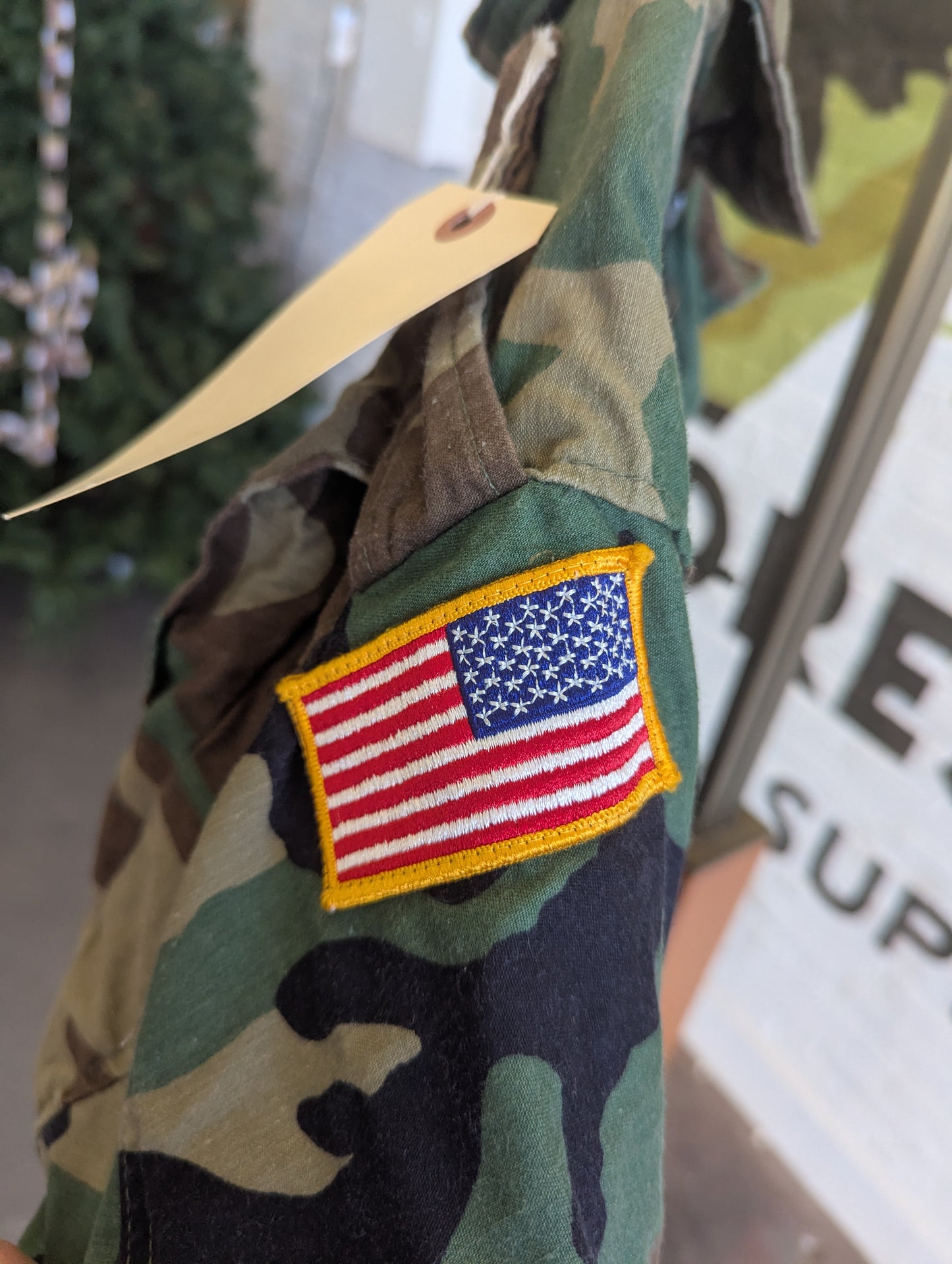 A close-up of a camouflage military-style jacket sleeve featuring a rectangular embroidered United States flag patch with a yellow border, set against a softly blurred indoor background.