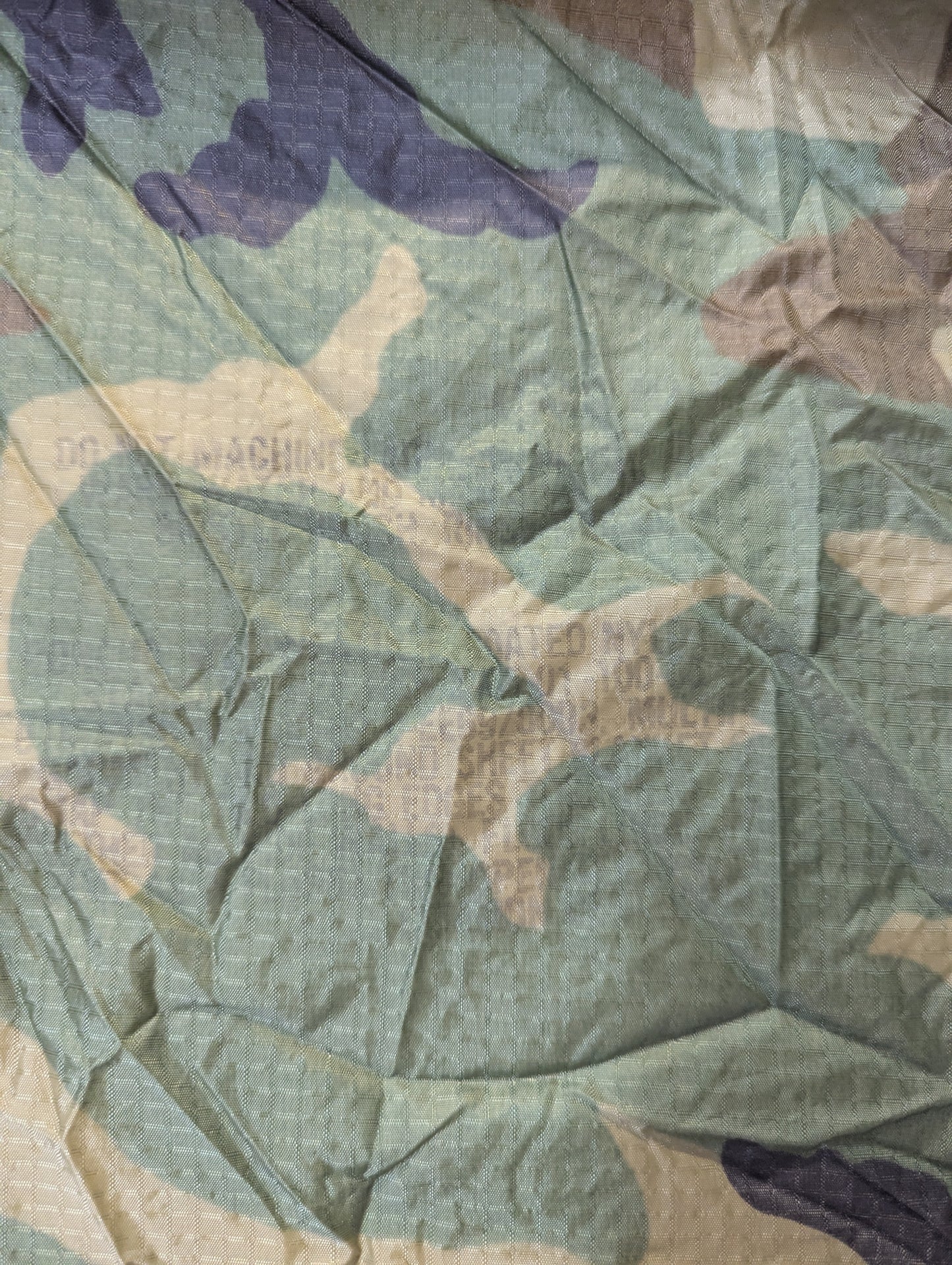 A close-up of a camouflage-patterned fabric with a crinkled texture, featuring shades of sage green, beige, brown, and dark purple.