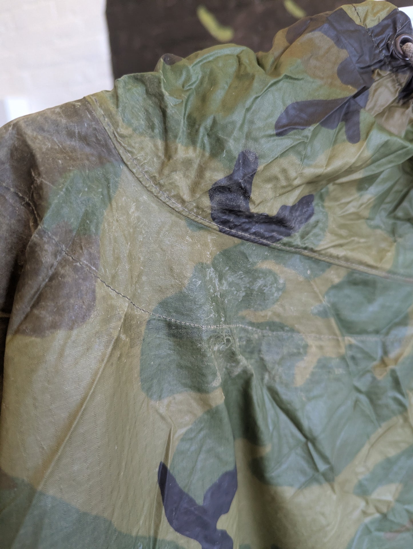 Close-up of a camouflage-patterned hooded rain jacket or poncho, showing the fabric texture, stitching, and a drawstring hood in olive green, tan, and dark gray.