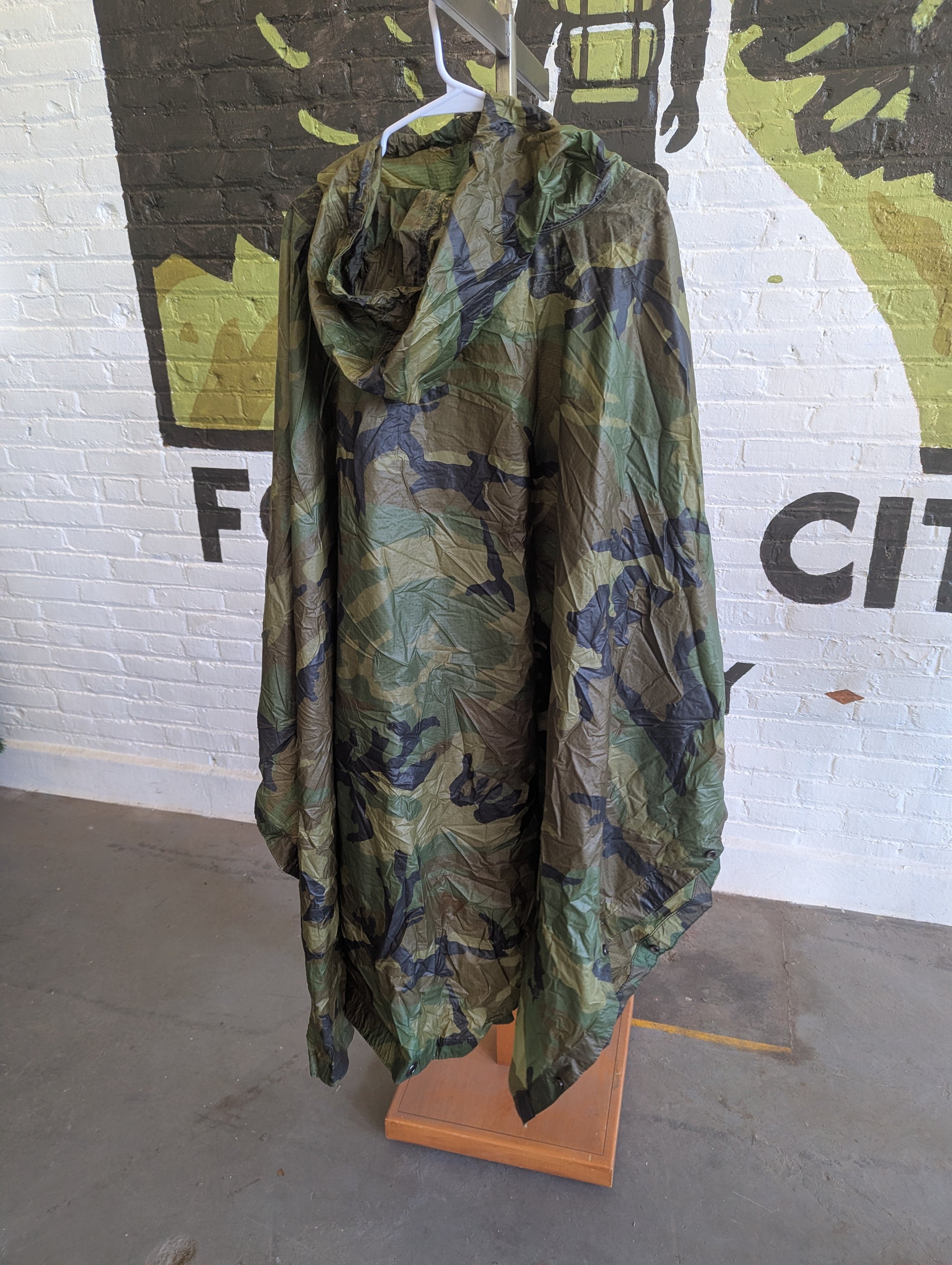 A hooded rain poncho with a green, brown, and black leafy camouflage pattern, hanging on a white hanger from a wooden stand in front of a white brick wall with graffiti.