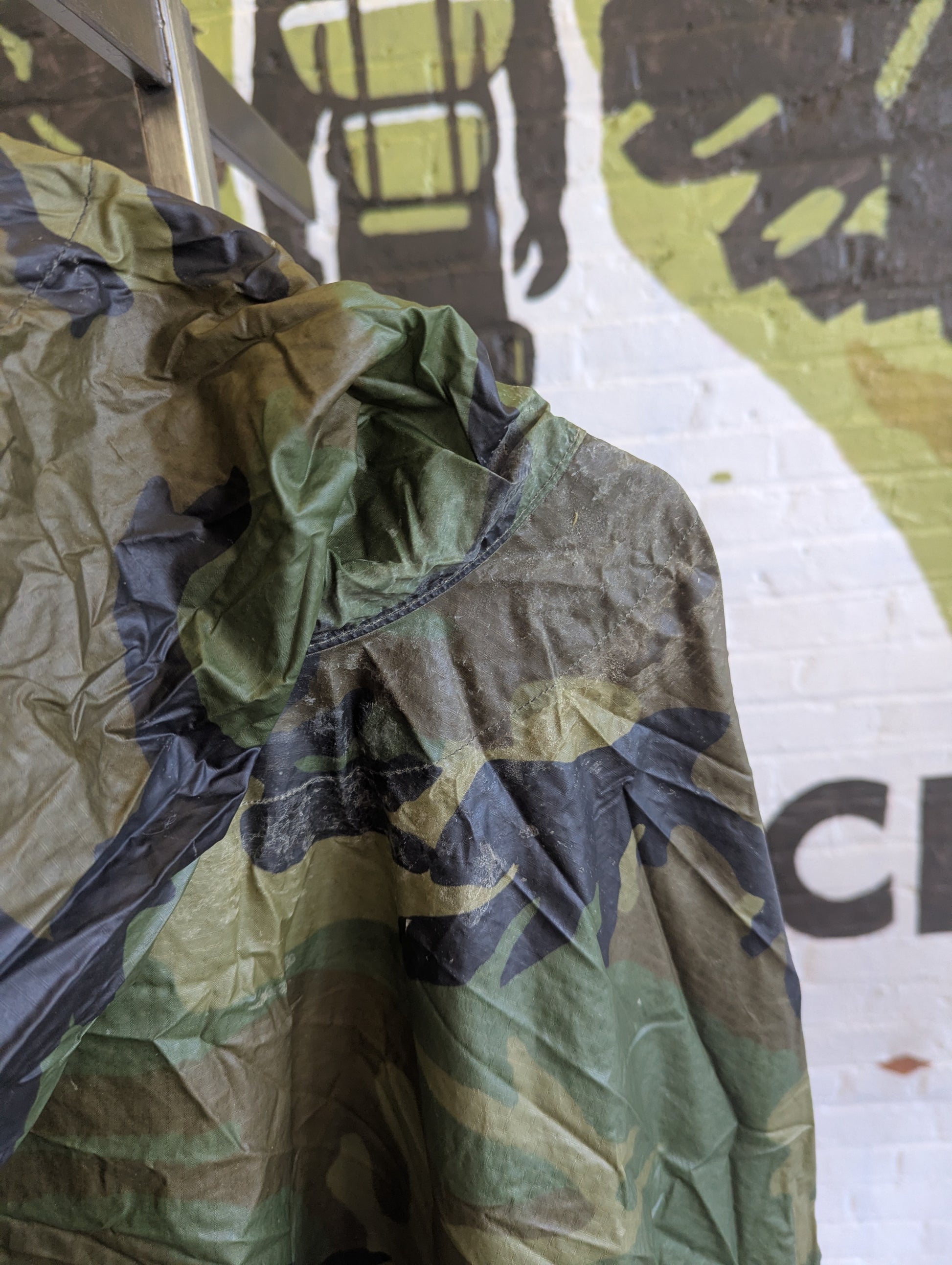 A camouflage hooded rain jacket with a green, brown, and black pattern, draped over a rack against a white brick wall with a graffiti mural.