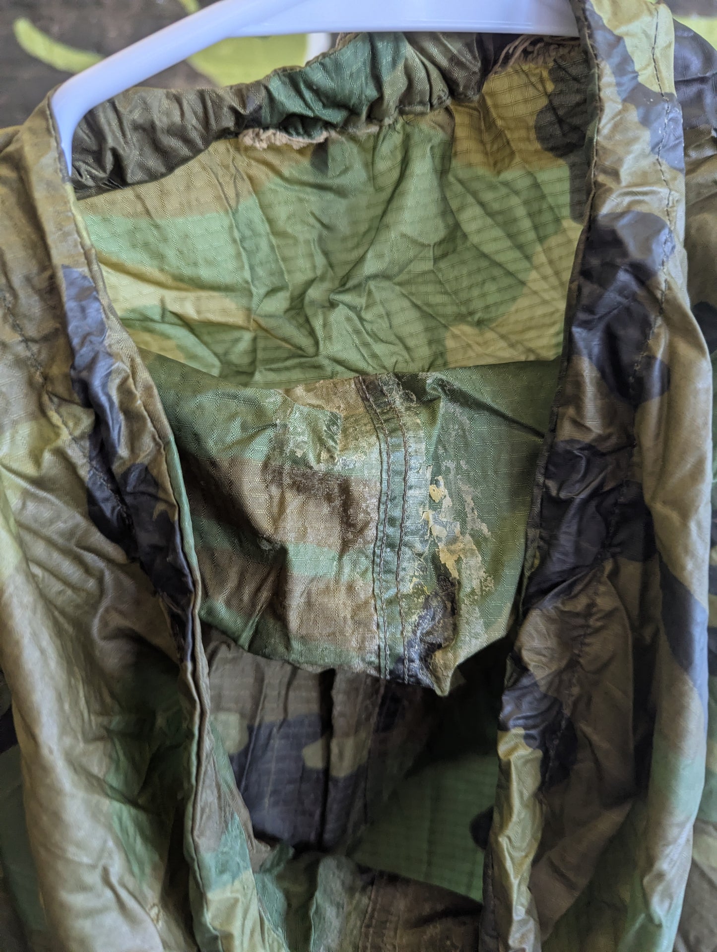 A camouflage military-style jacket or field gear hanging on a white plastic hanger, showing a wrinkled olive green and brown patterned fabric interior and outer shell.