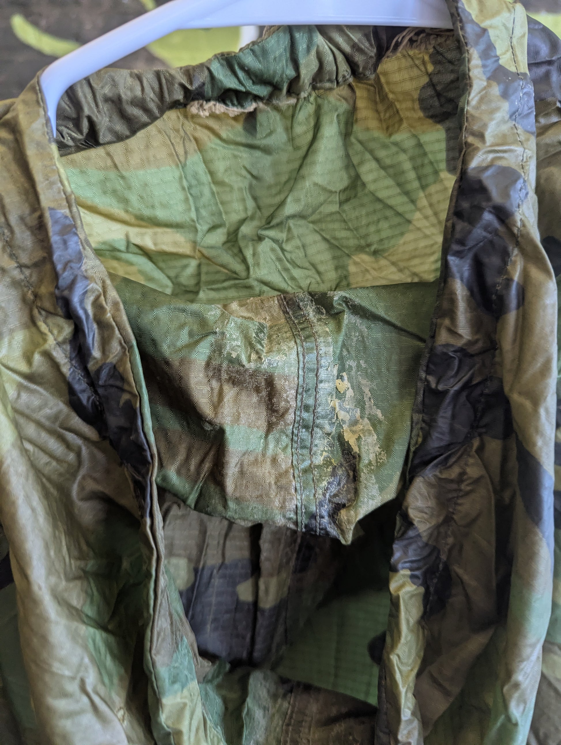 A camouflage military-style jacket or field gear hanging on a white plastic hanger, showing a wrinkled olive green and brown patterned fabric interior and outer shell.