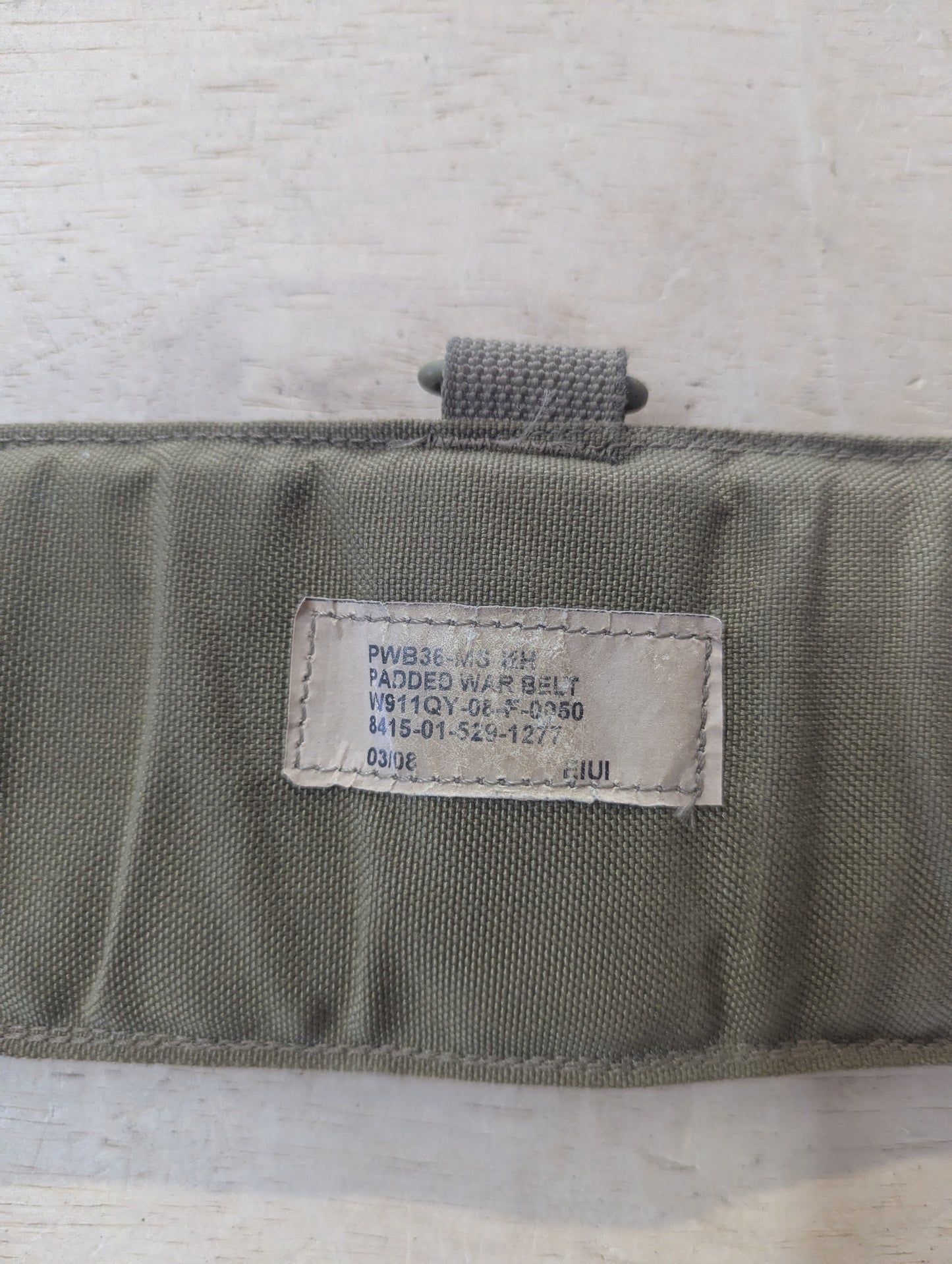 An olive drab padded war belt with a stitched fabric label reading 'PWB36 W/ KH PADDED WAR BELT' on a light wood background.