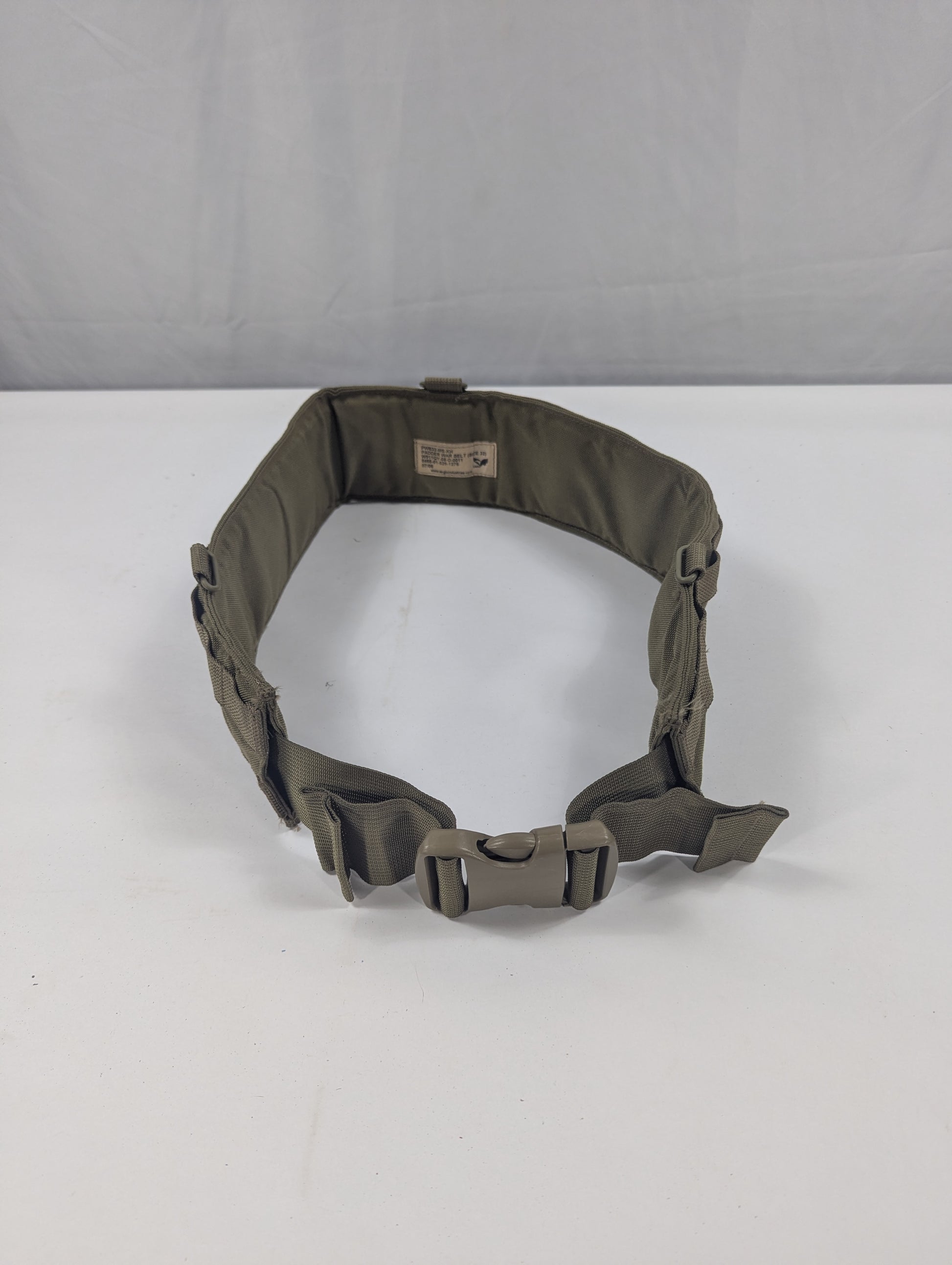 An olive drab tactical belt with a quick-release plastic buckle, displayed on a white circular stand against a white fabric backdrop.