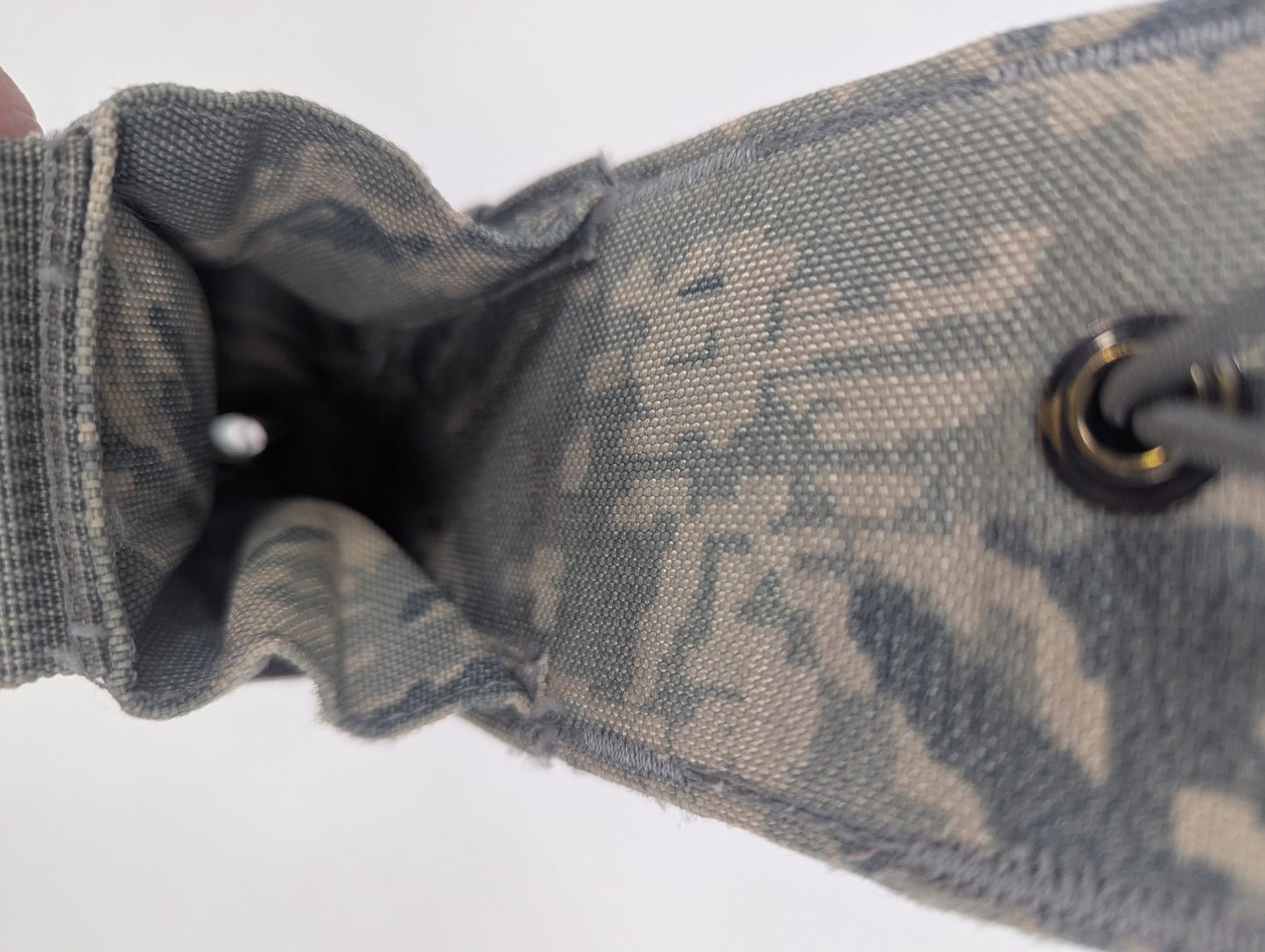 Close-up of a drawstring pouch or bag material in a gray and beige camouflage pattern, showing the fabric texture, interior opening, and a metal grommet on a white background.