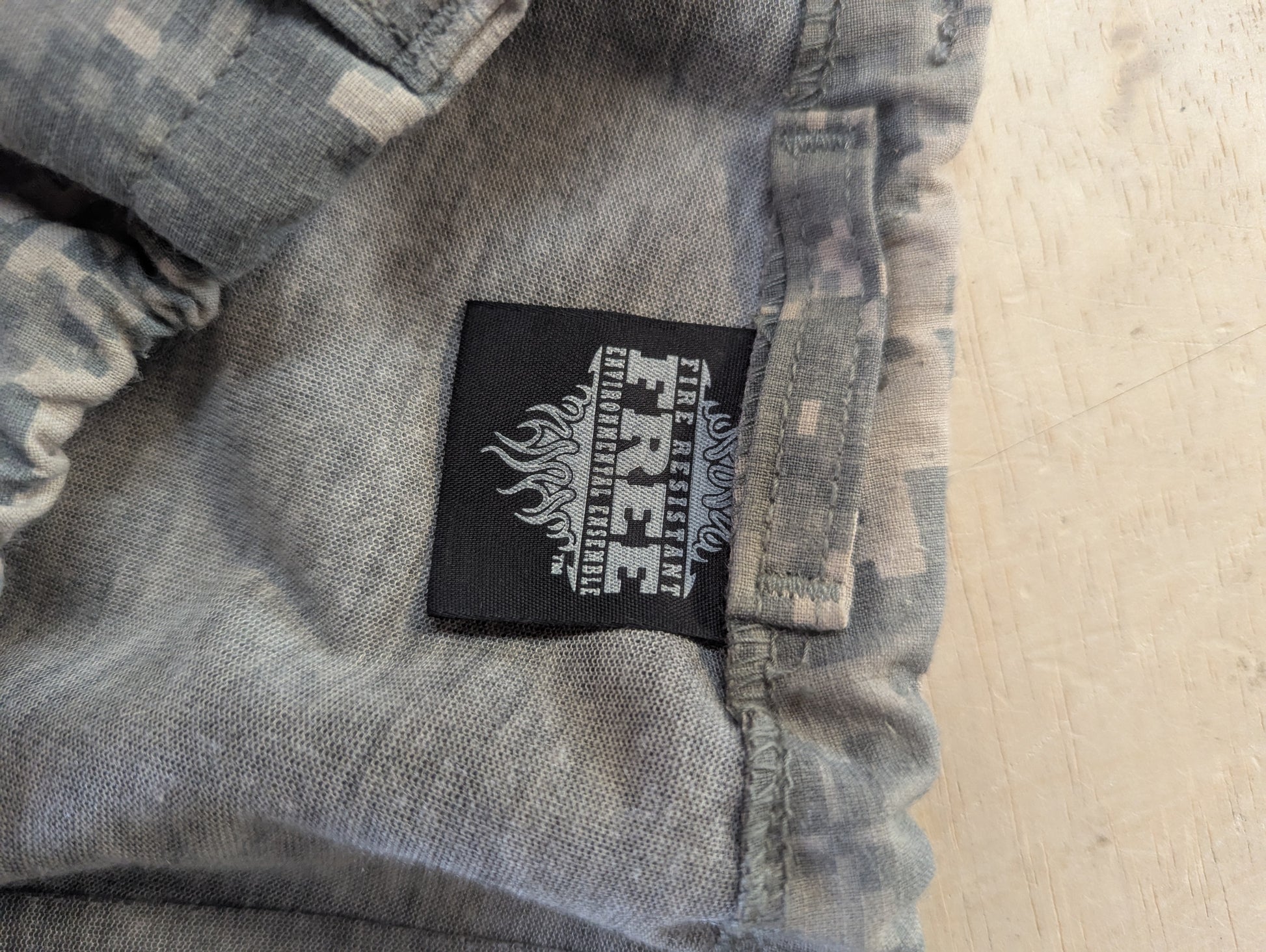 Close-up of a pair of gray camouflage pants showing a black rectangular brand patch with white text that reads 'FIRE RESCUE' and 'THE FIRE RESCUE COMMAND'.