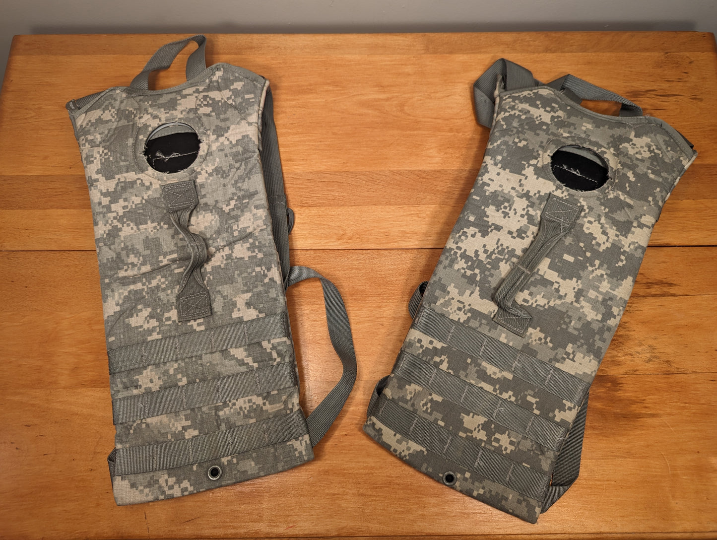 A pair of used U.S. Military Hydration Carriers in ACU digital camouflage, laid side-by-side on a wooden surface, displaying the MOLLE webbing, shoulder straps, and filler ports.