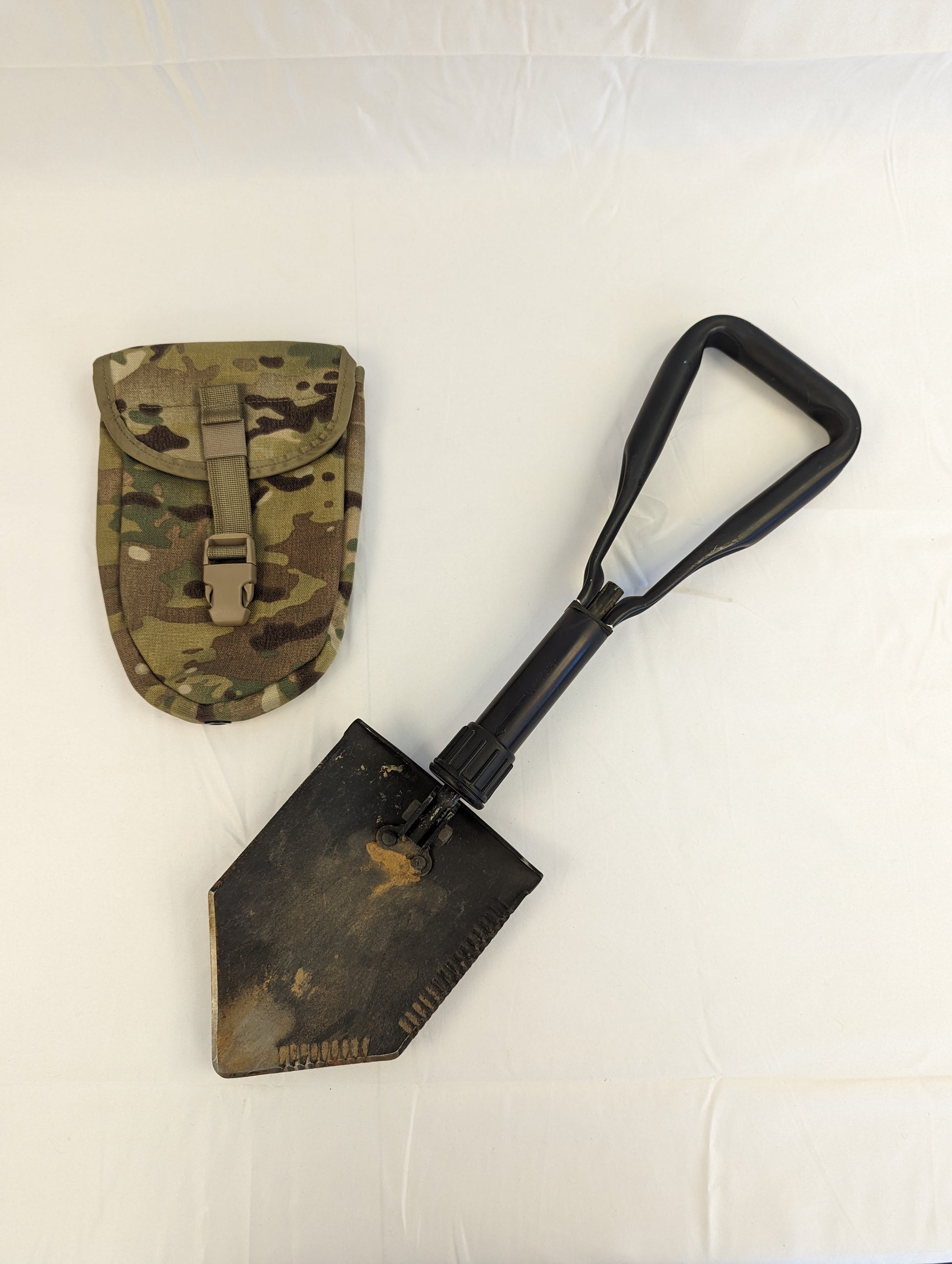 A used, black AMES Entrenching Tool with a soiled steel blade and a black plastic D-handle, resting next to its OCP/Multicam camouflage storage pouch on a white background.