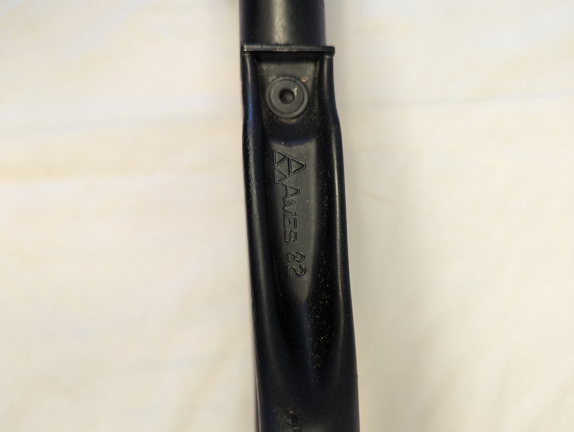 A close-up photograph of the black metal handle of the AMES Entrenching Tool, showing the embossed 'AMES 82' manufacturer's stamp and an attachment rivet.