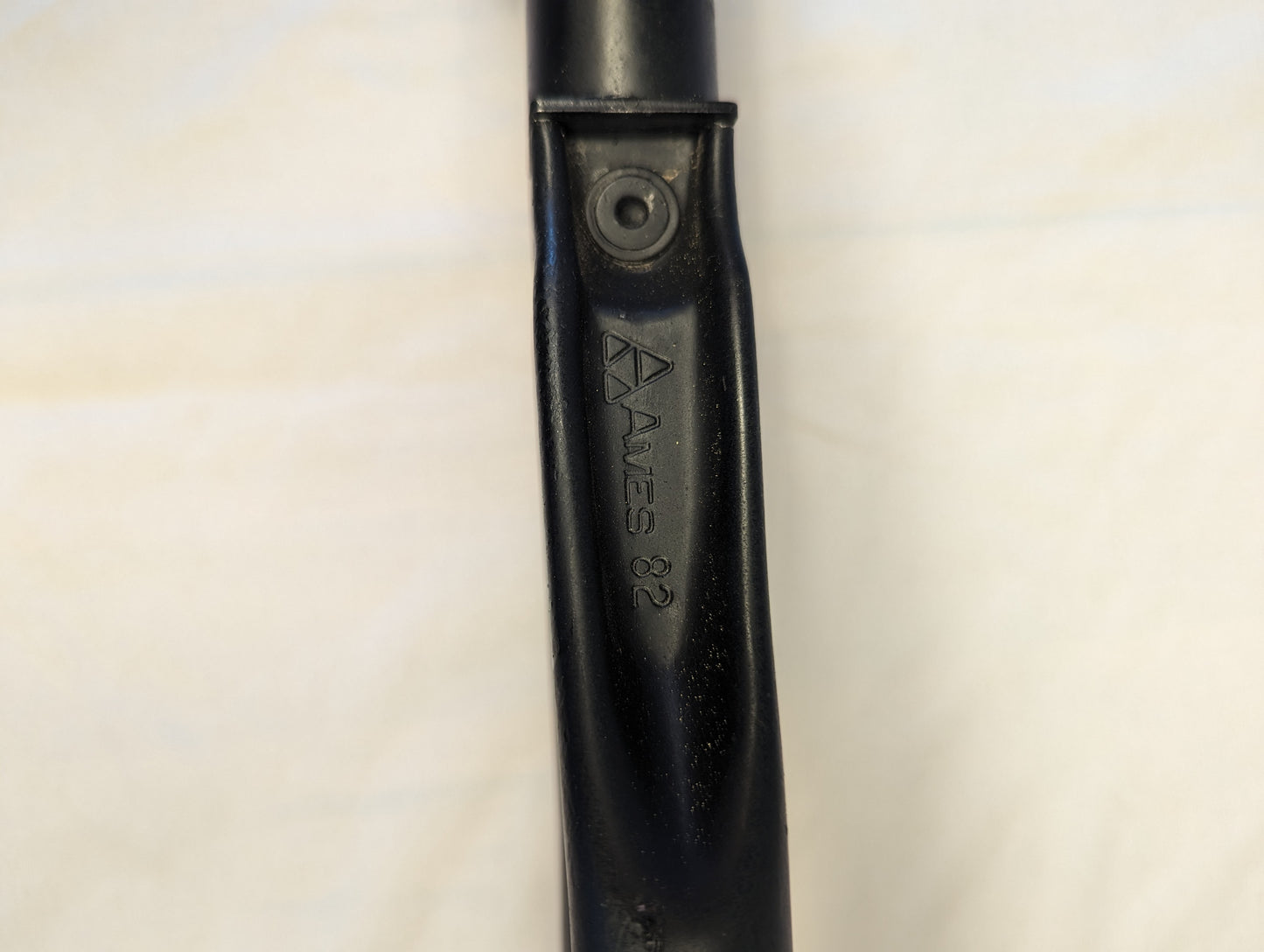 A close-up photograph of the black metal handle of the AMES Entrenching Tool, showing the embossed 'AMES 82' manufacturer's stamp and an attachment rivet.