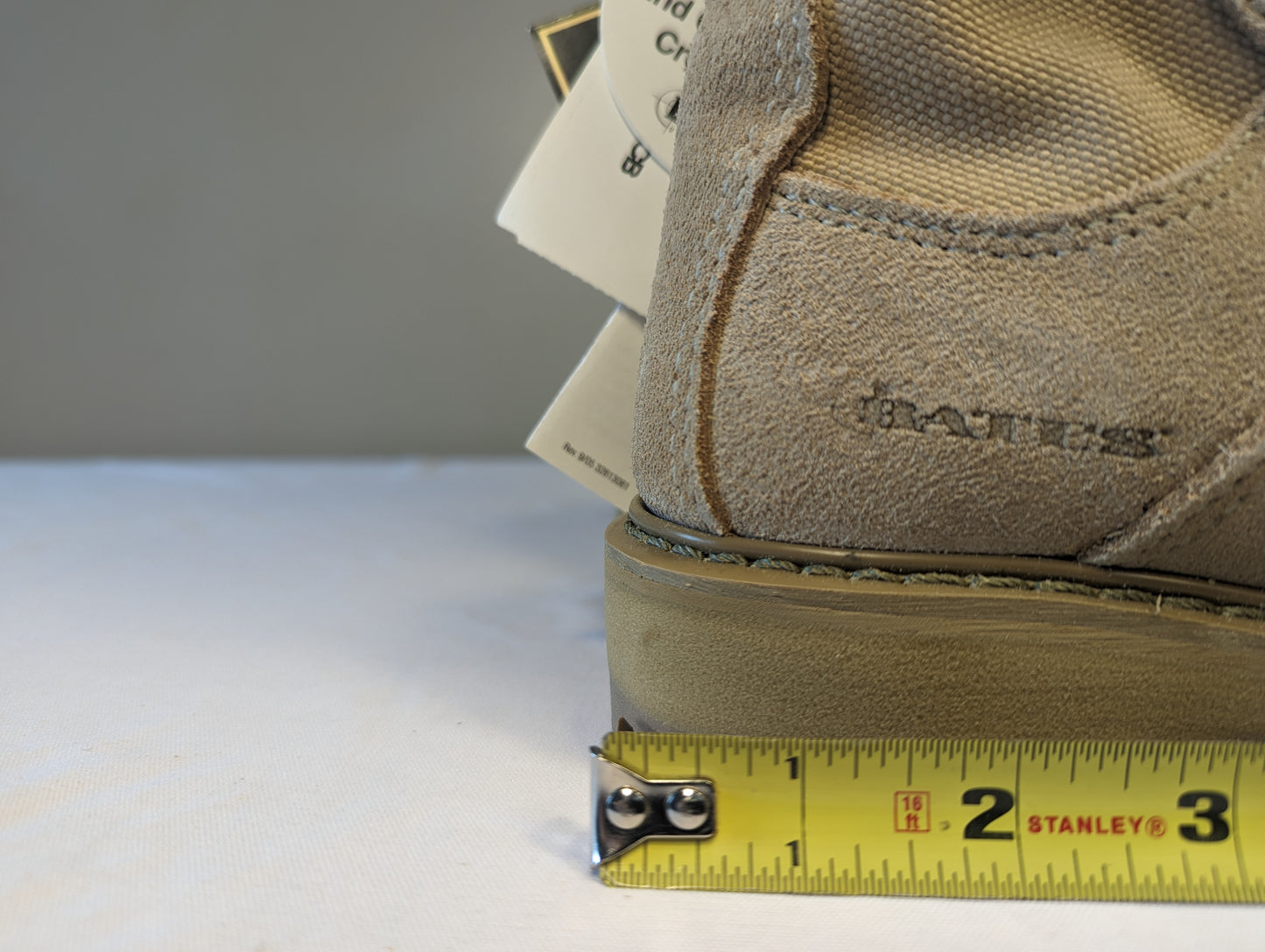 A close-up view of the heel of a tan Bates Cold Weather Combat Boot, displaying the embossed 'BATES' logo and a yellow Stanley tape measure demonstrating the sole thickness and heel width measurement.