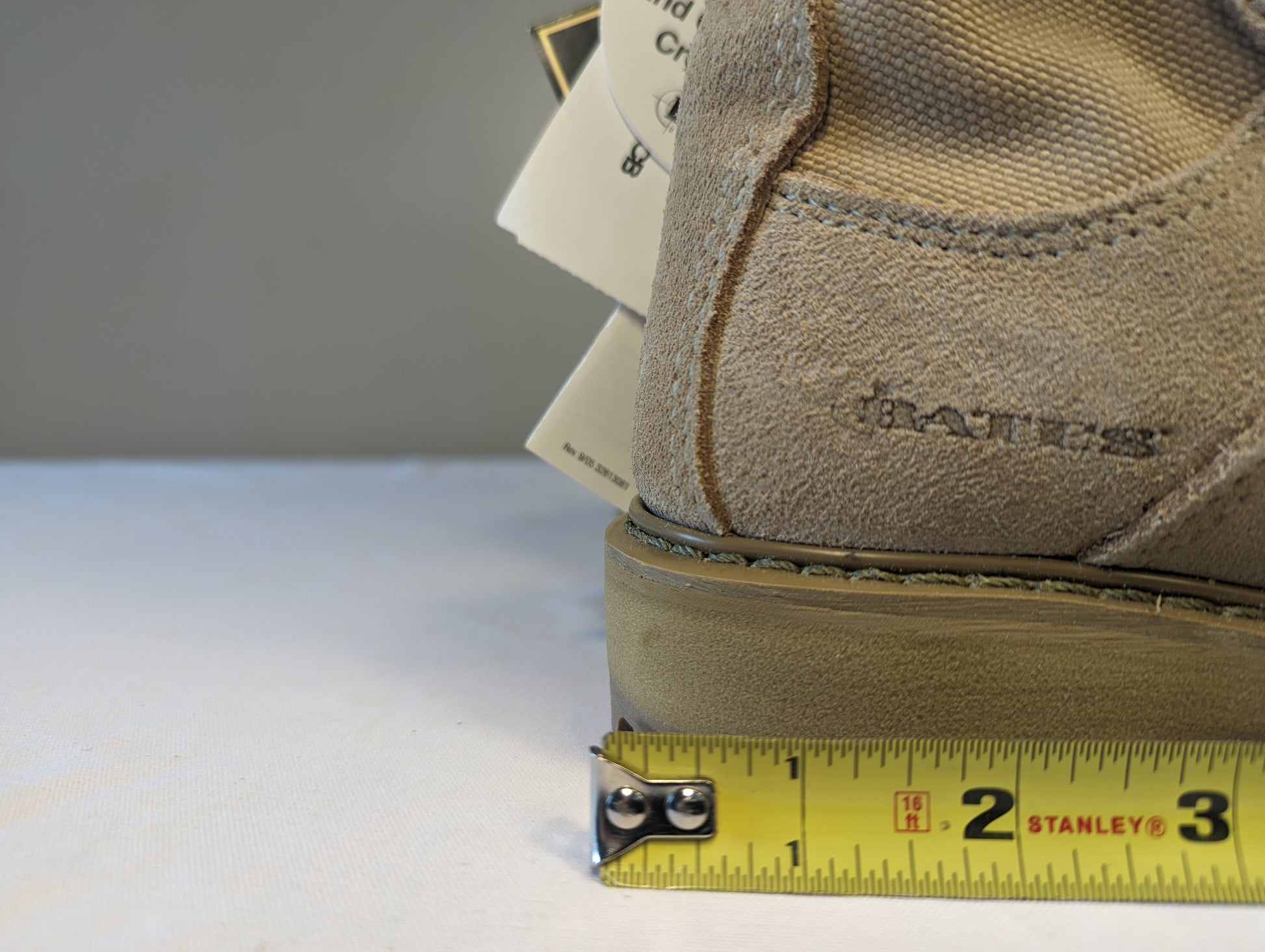 A close-up view of the heel of a tan Bates Cold Weather Combat Boot, displaying the embossed 'BATES' logo and a yellow Stanley tape measure demonstrating the sole thickness and heel width measurement.