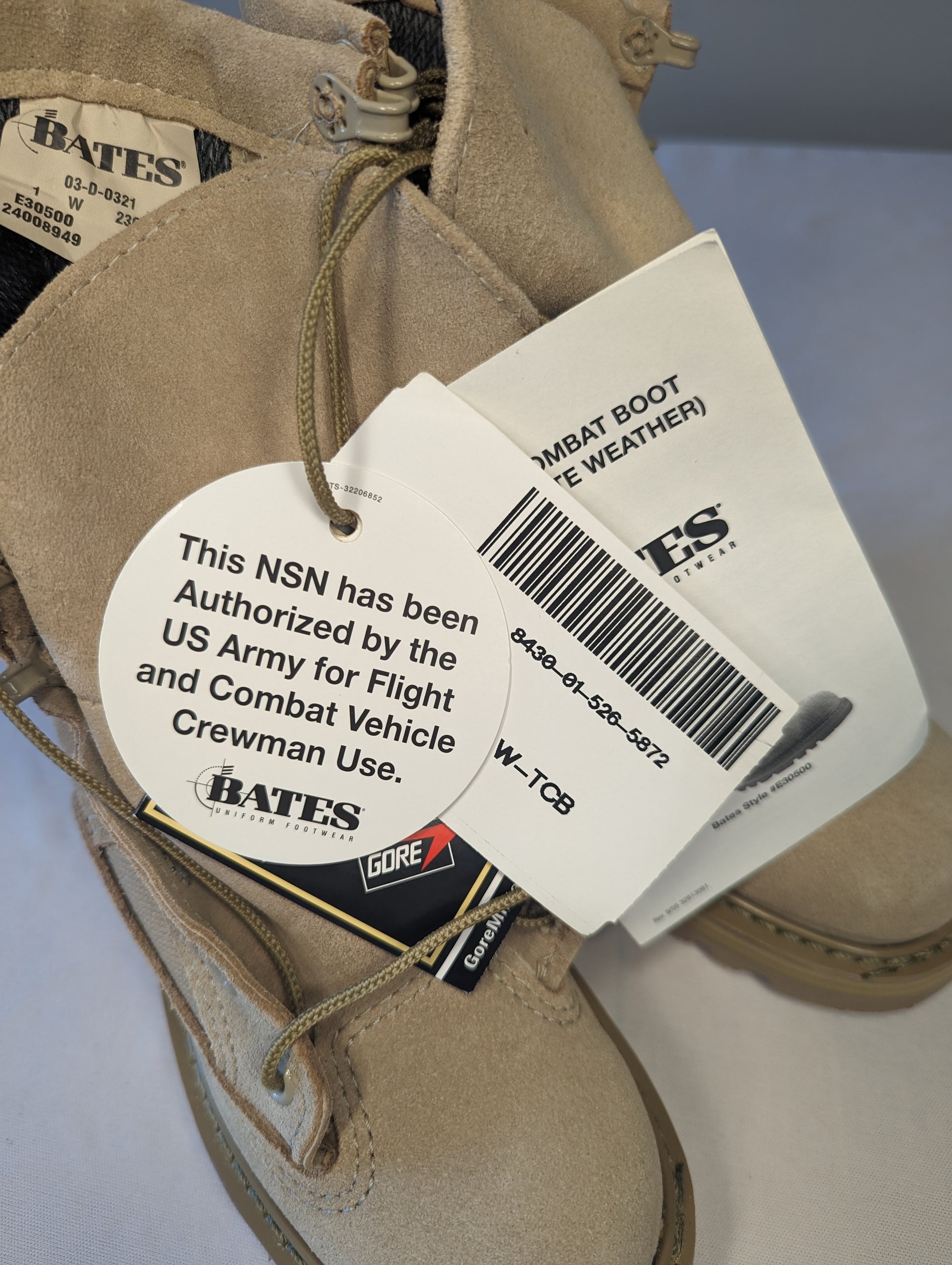 A close-up of a tan Bates Cold Weather Combat Boot, displaying the official U.S. Army hang tag which authorizes the boot for Flight and Combat Vehicle Crewman use, along with the Gore-Tex tag.