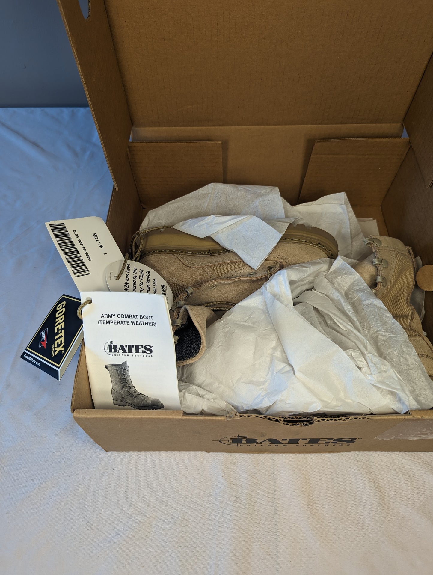 An overhead view into an open cardboard box, showing a new tan Bates Cold Weather Combat Boot nestled in tissue paper, with the BATES logo stamped inside the box lid and the official instruction tag visible.