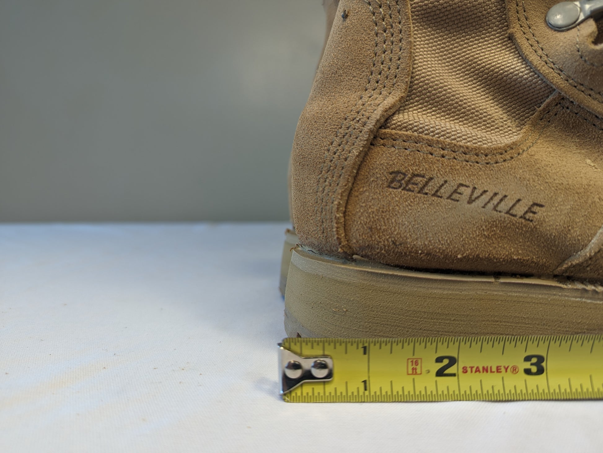 A close-up view of the heel of a tan Belleville Cold Weather Combat Boot, displaying the embossed 'BELLEVILLE' logo and a yellow tape measure demonstrating the sole thickness and heel width.